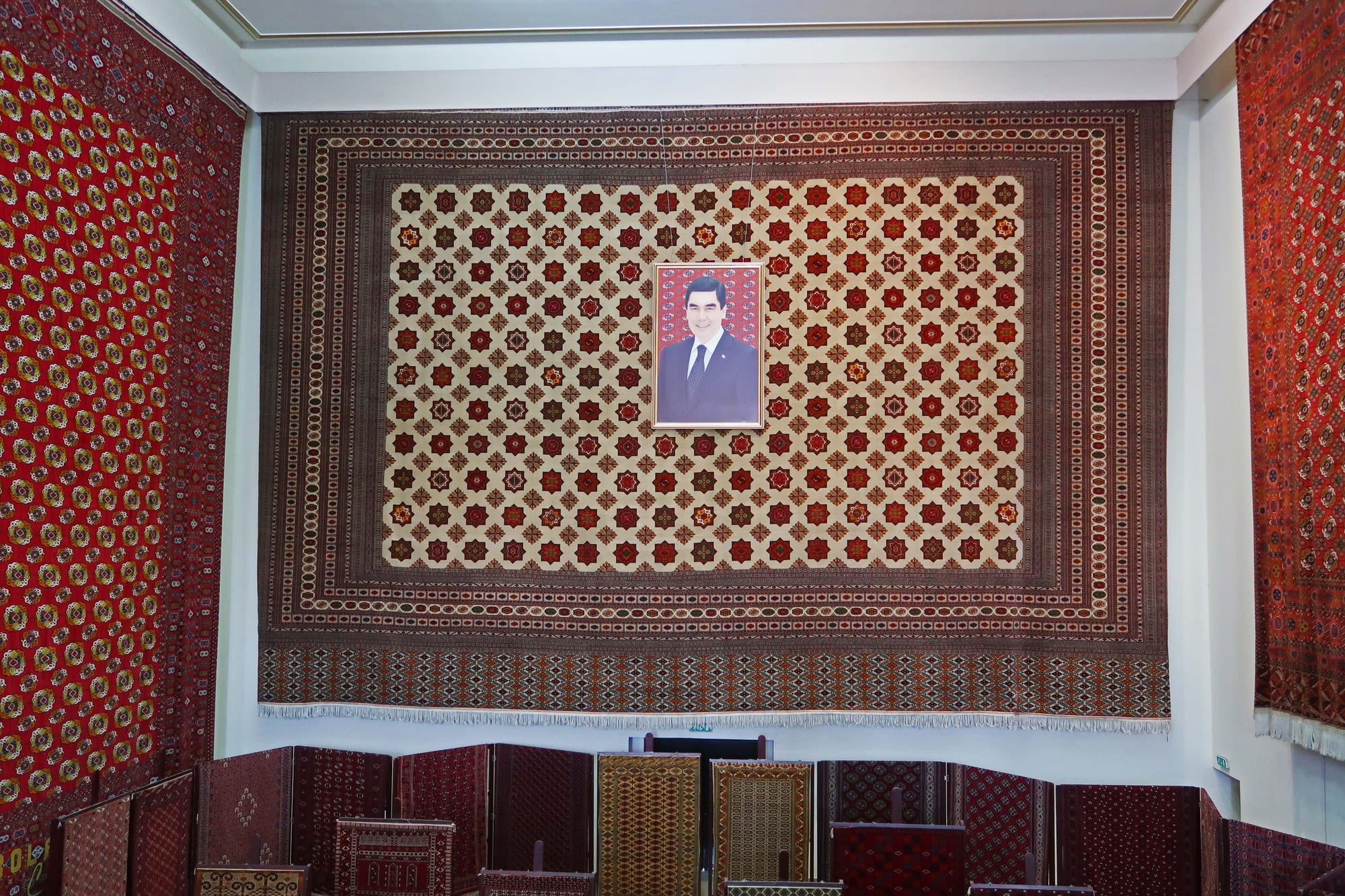 A museum hall hung with large red Turkmen carpets patterned with repeating medallions, with a framed portrait of a man in a suit at the centre of the largest carpet and smaller rugs displayed below