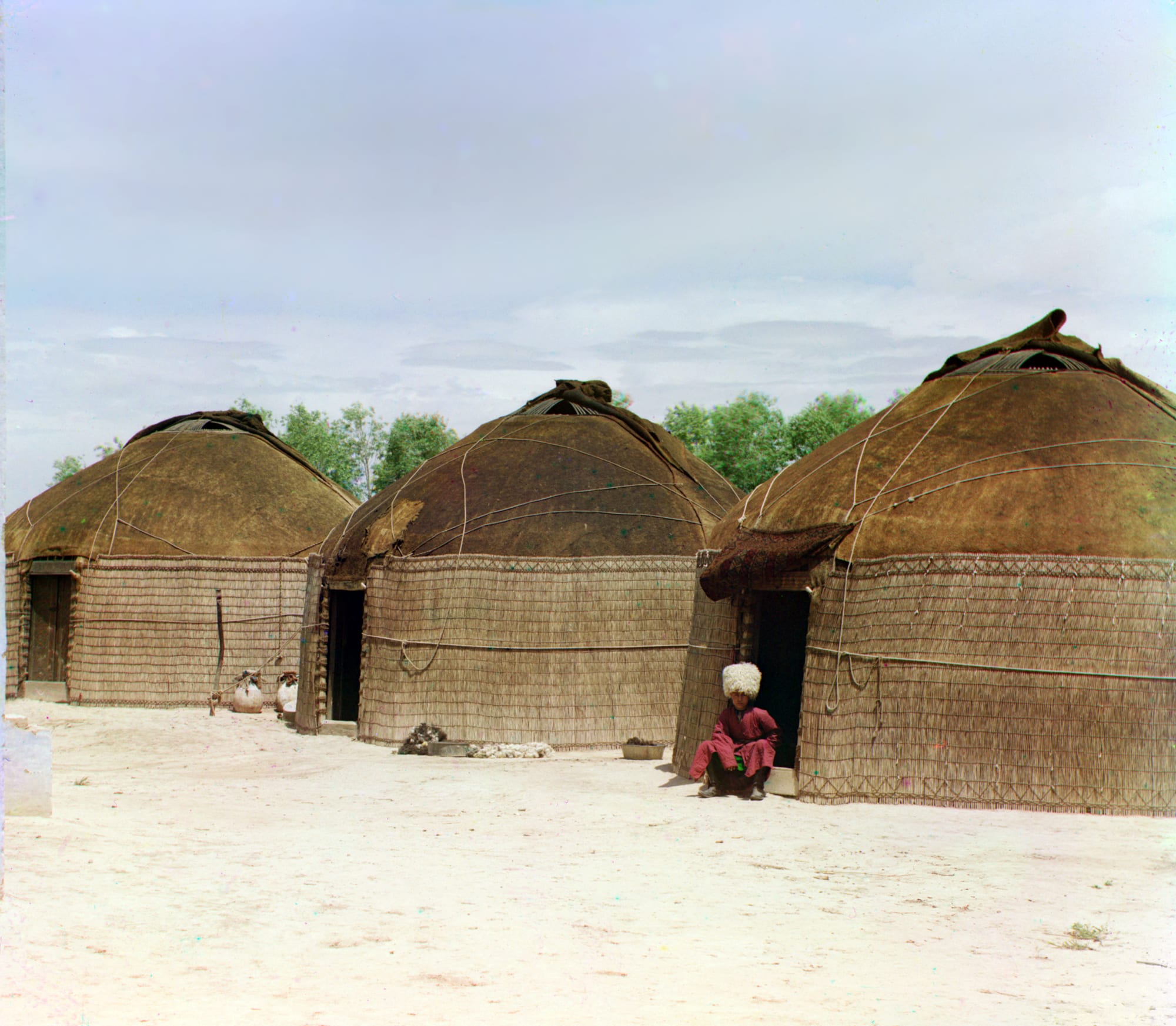 An early colour photograph of three round, dome-roofed yurts with reed-mat walls on pale bare ground, a person in red sitting at one doorway