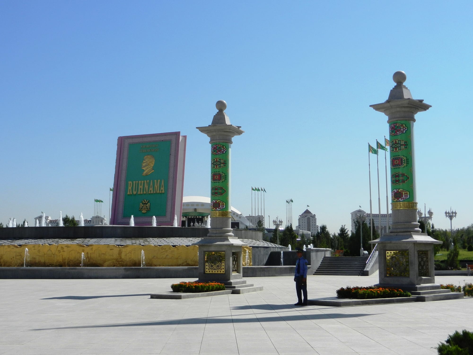 A giant pink-and-green monument shaped like a closed book lettered Ruhnama with a gold profile portrait, behind fountains and two tall columns decorated with carpet patterns on a wide paved square