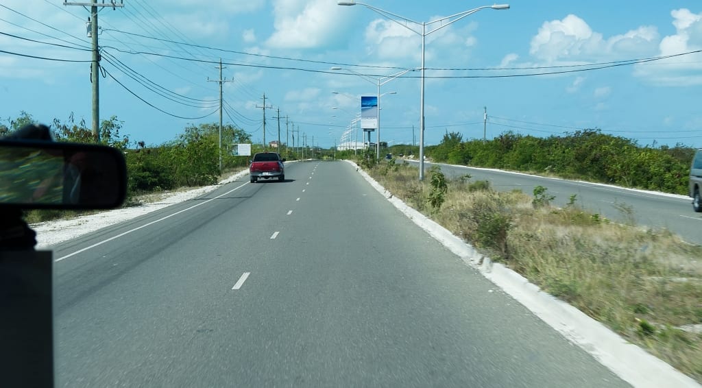 A quiet two-lane highway lined with low scrub and palm trees on Providenciales, with a single car ahead and street lighting overhead