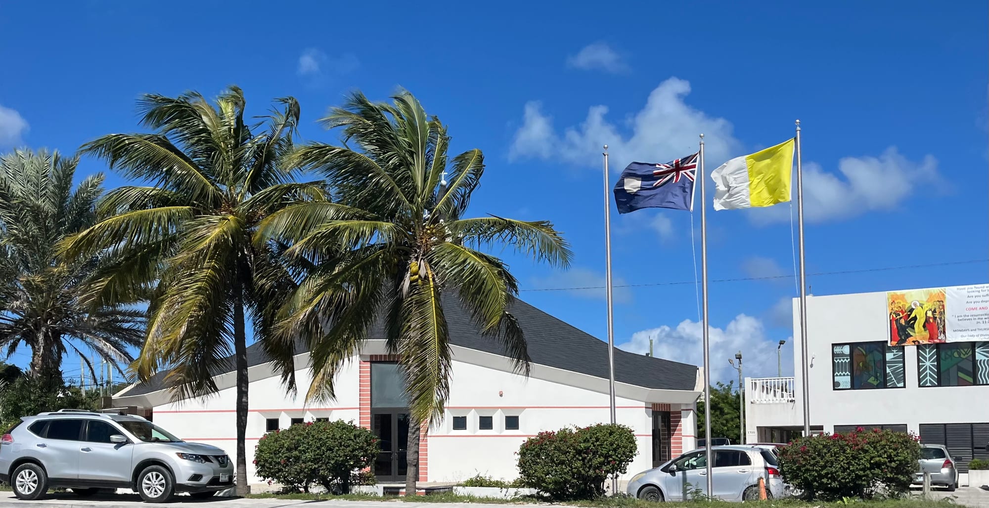 A low white church building with a sweeping roofline, palm trees, flagpoles and parked cars outside, under a bright blue sky
