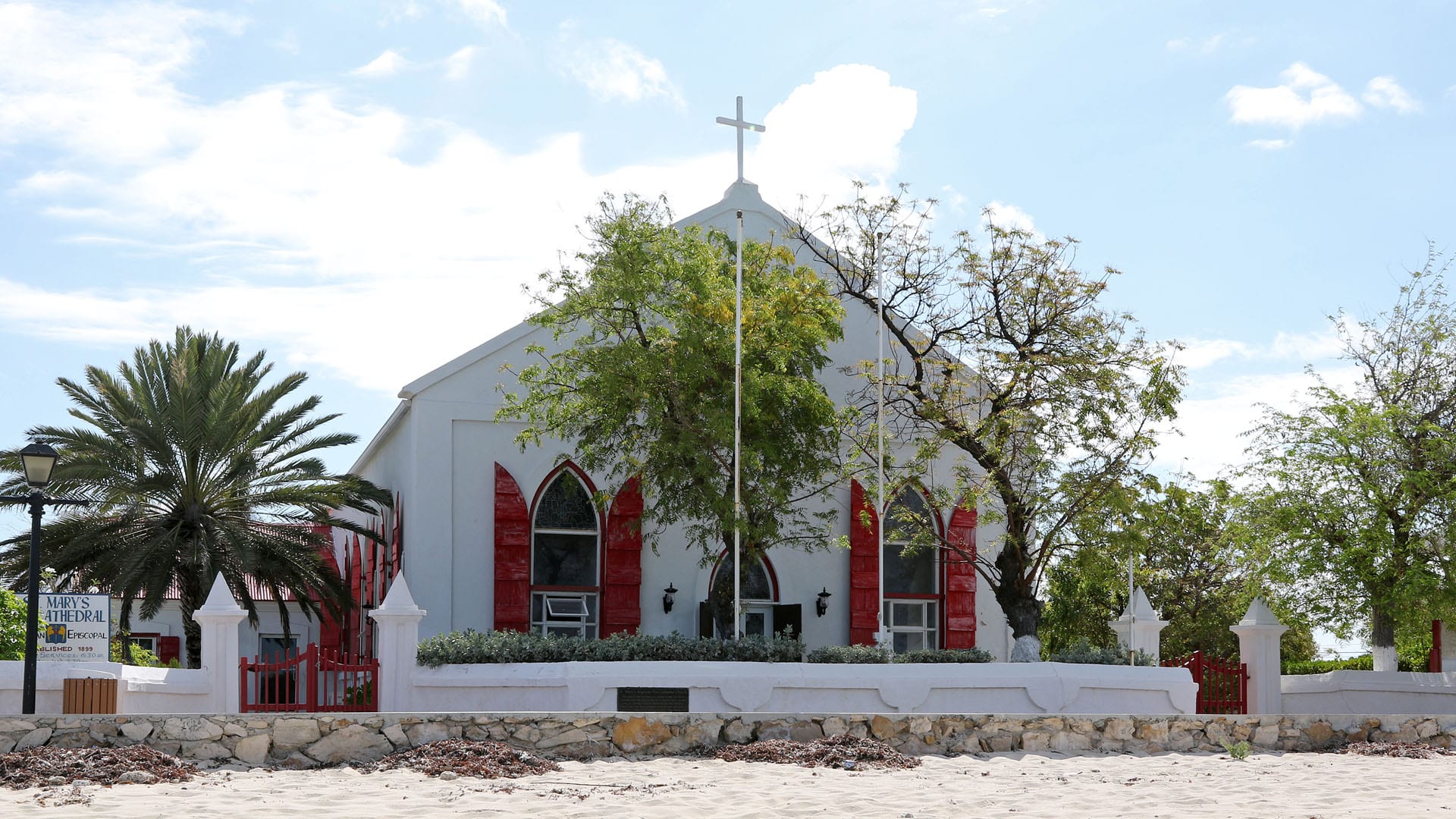 A white gabled church with a cross on the roof and bright red shutters, behind a low white wall with red gates, facing a sandy foreshore under a pale blue sky