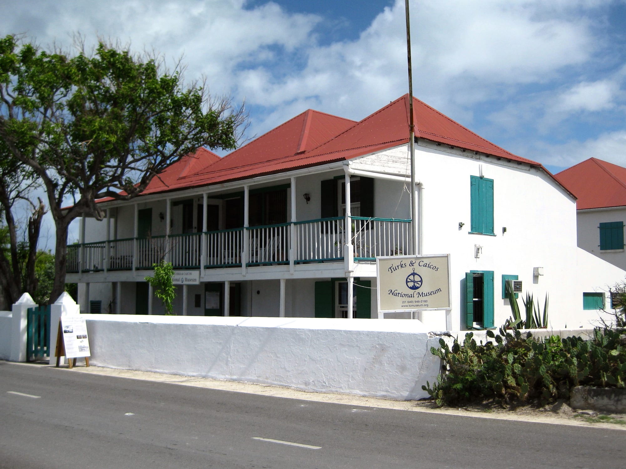 A white two-storey colonial house with a red roof, green shutters and a long first-floor balcony, behind a low white wall beside a road, with a sign reading Turks and Caicos National Museum