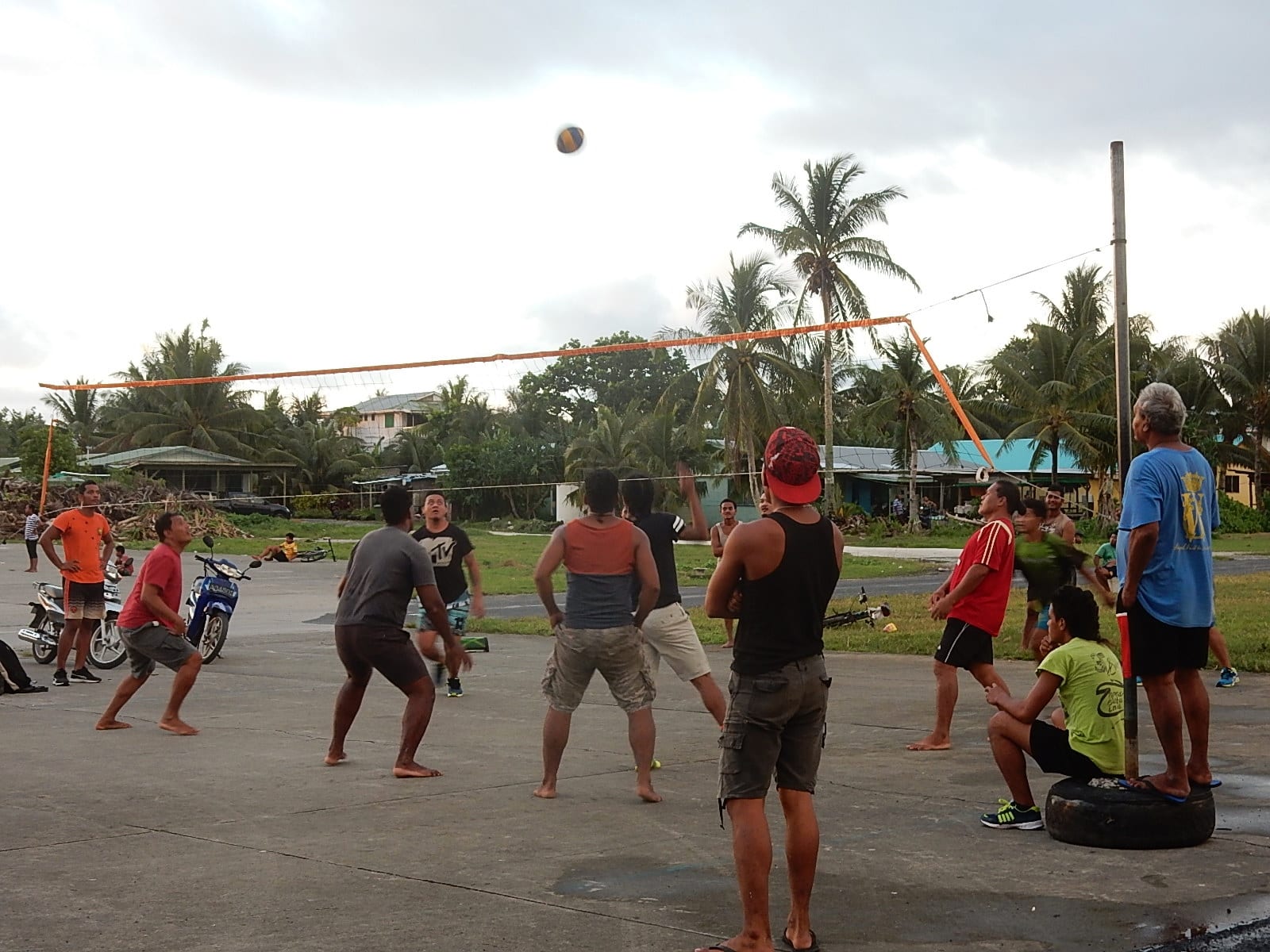 A group of men playing volleyball on the open tarmac of Funafuti's airport runway in the evening light, with palm trees and houses behind