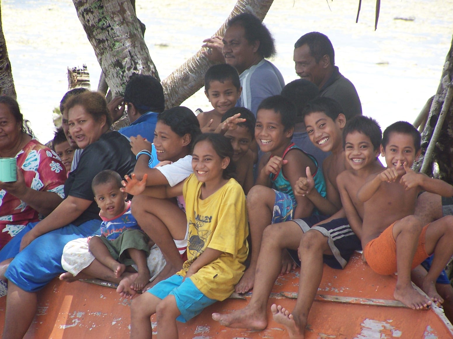 A large group of smiling adults and children, including several barefoot boys, sitting together on an orange boat hull under trees by the lagoon