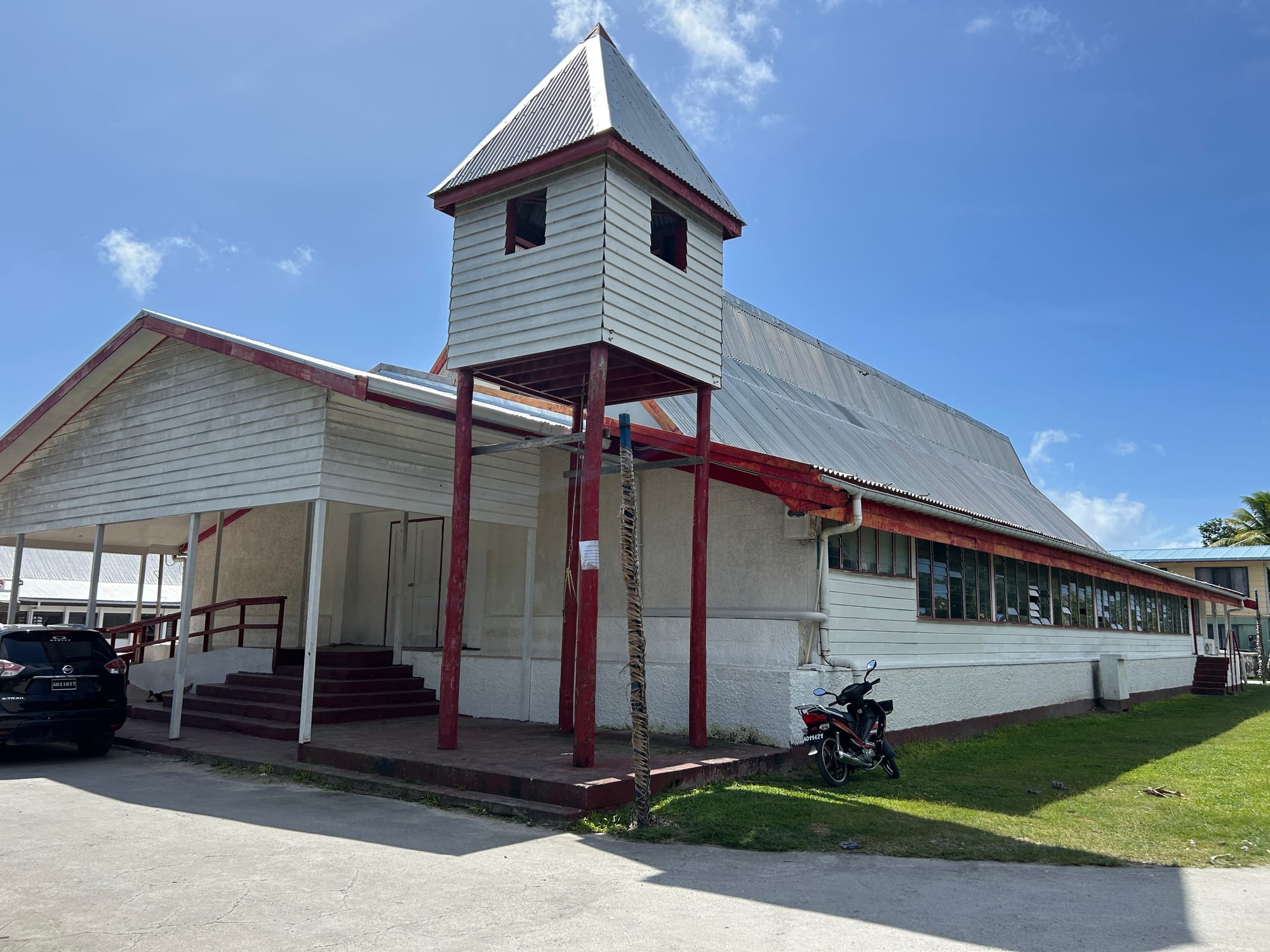 A white weatherboard church with red trim, a long corrugated metal roof and a boxy bell tower raised on red posts beside the entrance steps