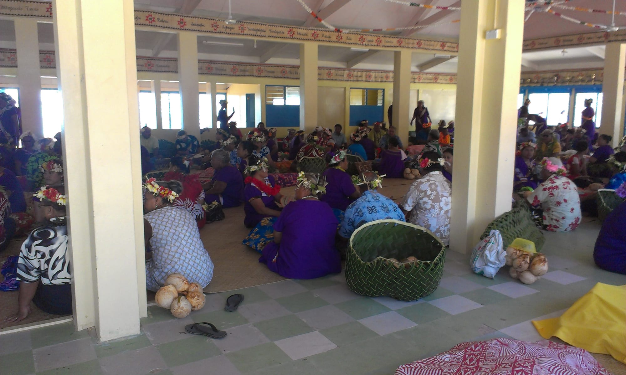 Women wearing flower garlands sit on mats on the floor of a large meeting hall, with woven green coconut-frond baskets and husked drinking coconuts set down among them