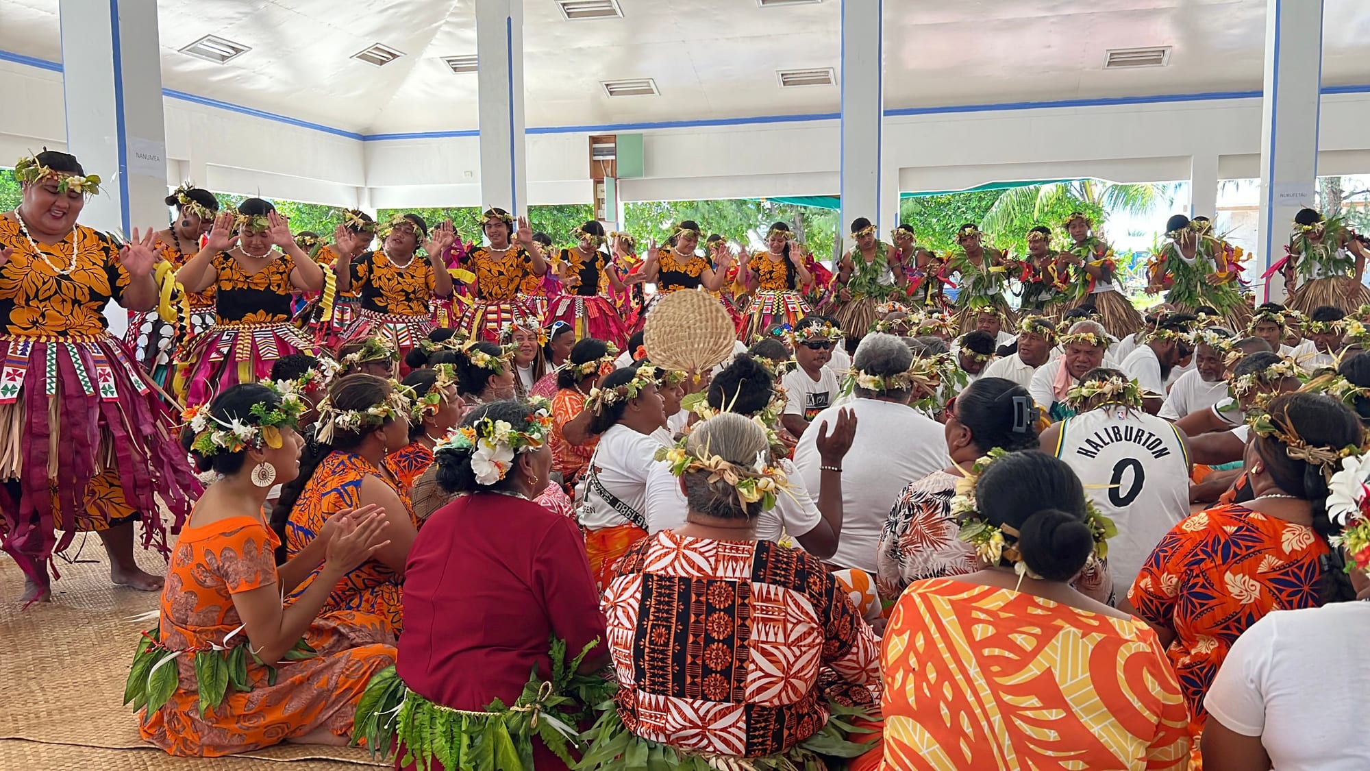 Women in orange and pink dance costumes and flower garlands dance with raised hands while a large group of garlanded singers sits on mats in front of them inside an open meeting hall