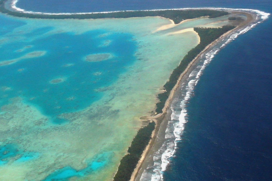 An aerial photo of Funafuti showing a thin curving strip of land with houses, the airstrip, and ocean on both sides