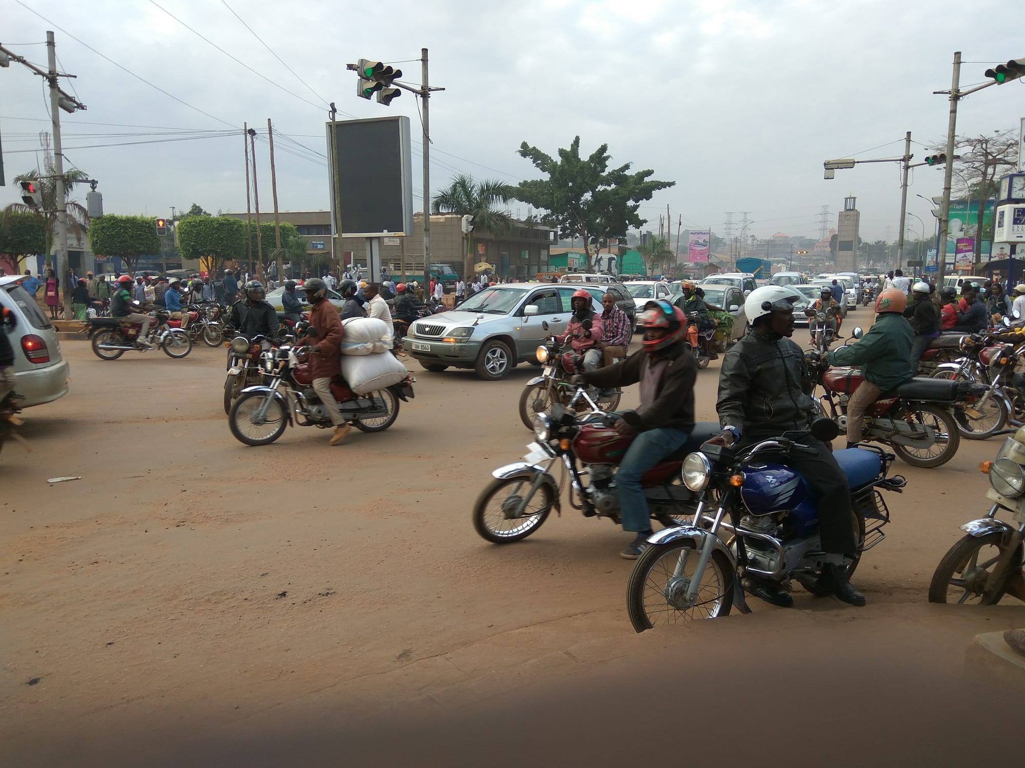 A busy Kampala intersection crowded with motorcycle taxis, cars and pedestrians under traffic lights, with dozens of boda-bodas queued at a junction