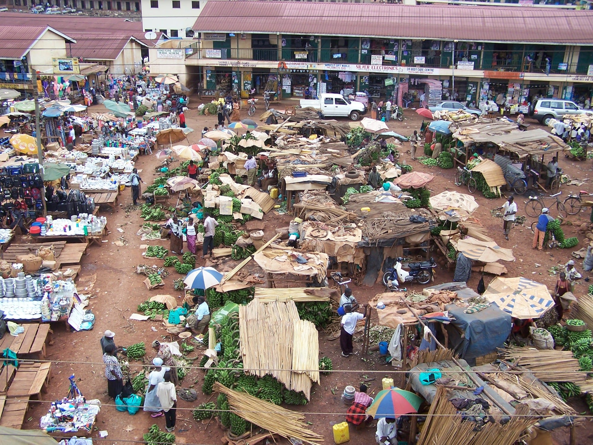 An aerial view of a bustling open-air Kampala market, with rows of stalls, umbrellas, banana bunches, general goods and dozens of shoppers moving through packed dirt lanes