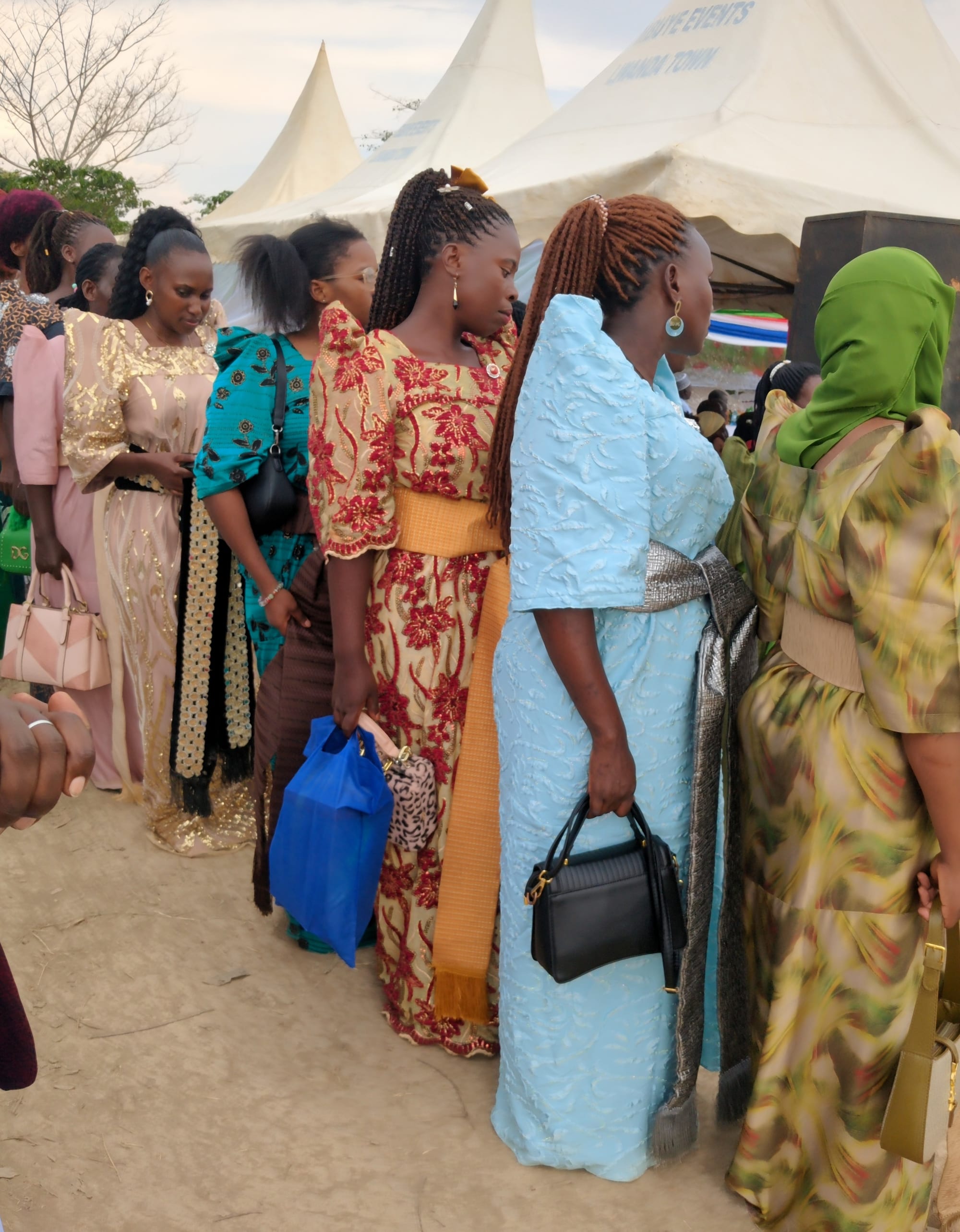 A line of women in brightly coloured gomesi dresses with wide sashes at the waist stand on a dirt path beside white event tents, seen partly from behind