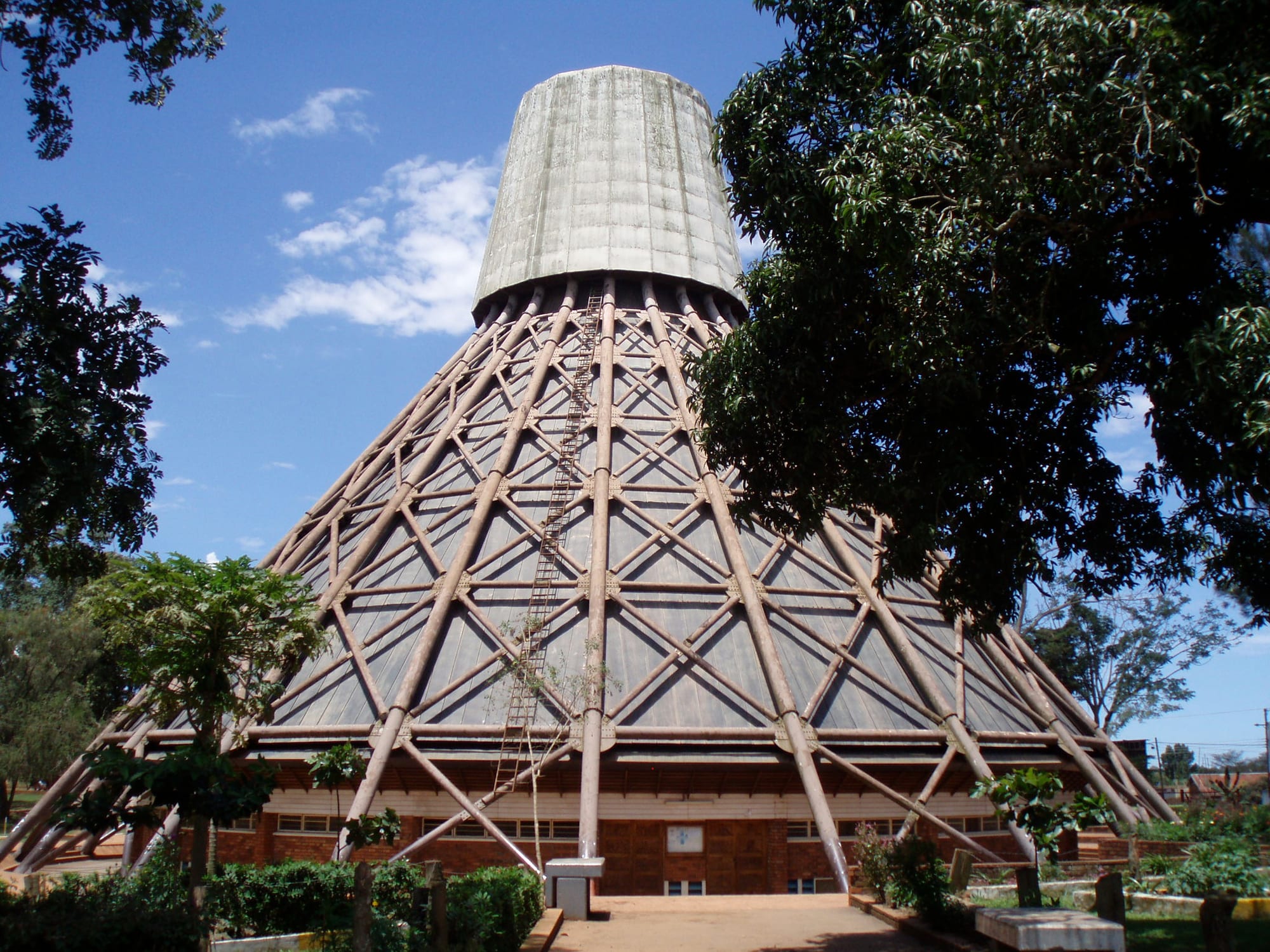A tall conical shrine building with a framework of long diagonal supports and a cylindrical top, set among trees under a blue sky