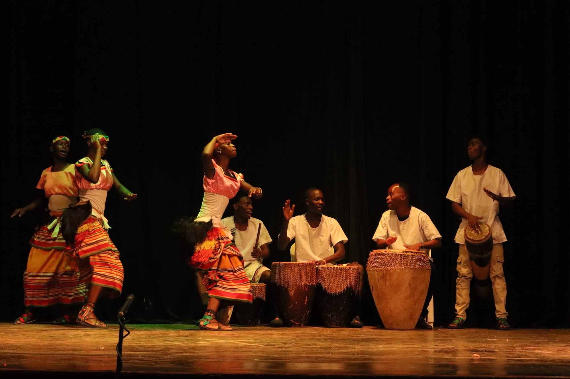 Three women in striped skirts and fringed hip wraps dance on a stage beside a row of large drums played by men in white shirts, against a dark curtain