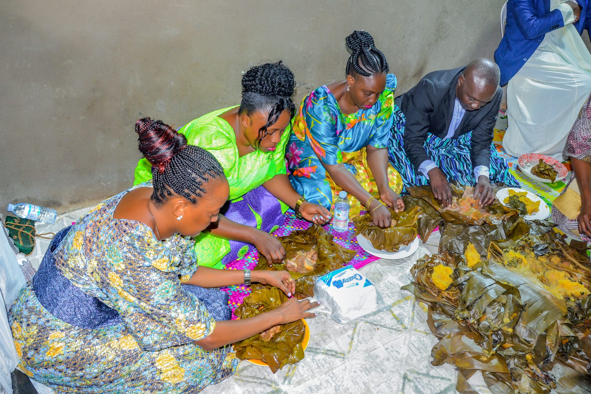 Three women in colourful gomesi and a man in a suit sit on a mat on the floor, eating with their hands from food wrapped in banana leaves on plates in front of them