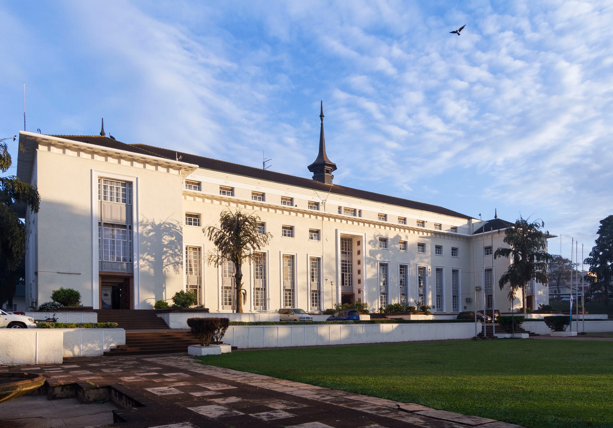 A long white three-storey building with a dark roof and a central spire, set behind a lawn and paved forecourt under a blue sky with thin cloud