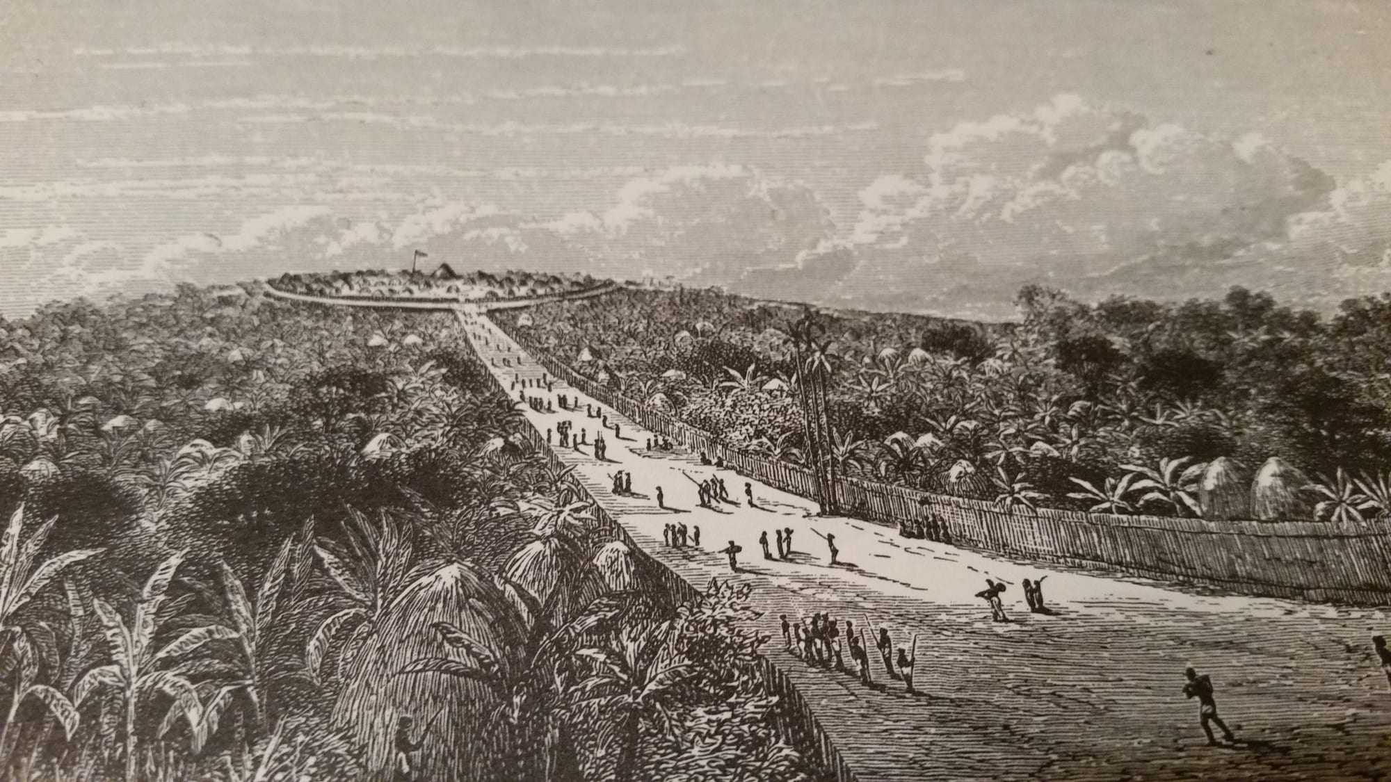 An old engraving of a broad fenced avenue running between banana gardens and thatched houses up to a hilltop enclosure with a flag, with small groups of people walking along it