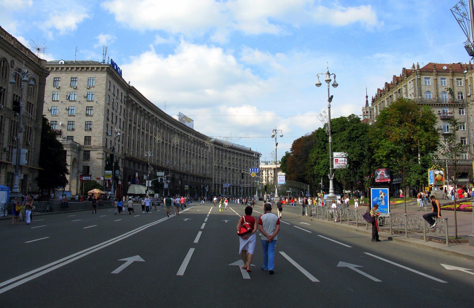 A wide, tree-lined boulevard in central Kyiv closed to traffic, with people walking and a young man skateboarding along painted road markings