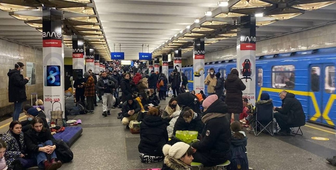 Dozens of people, including children, sitting and standing with luggage on a crowded Kyiv metro platform as a train arrives