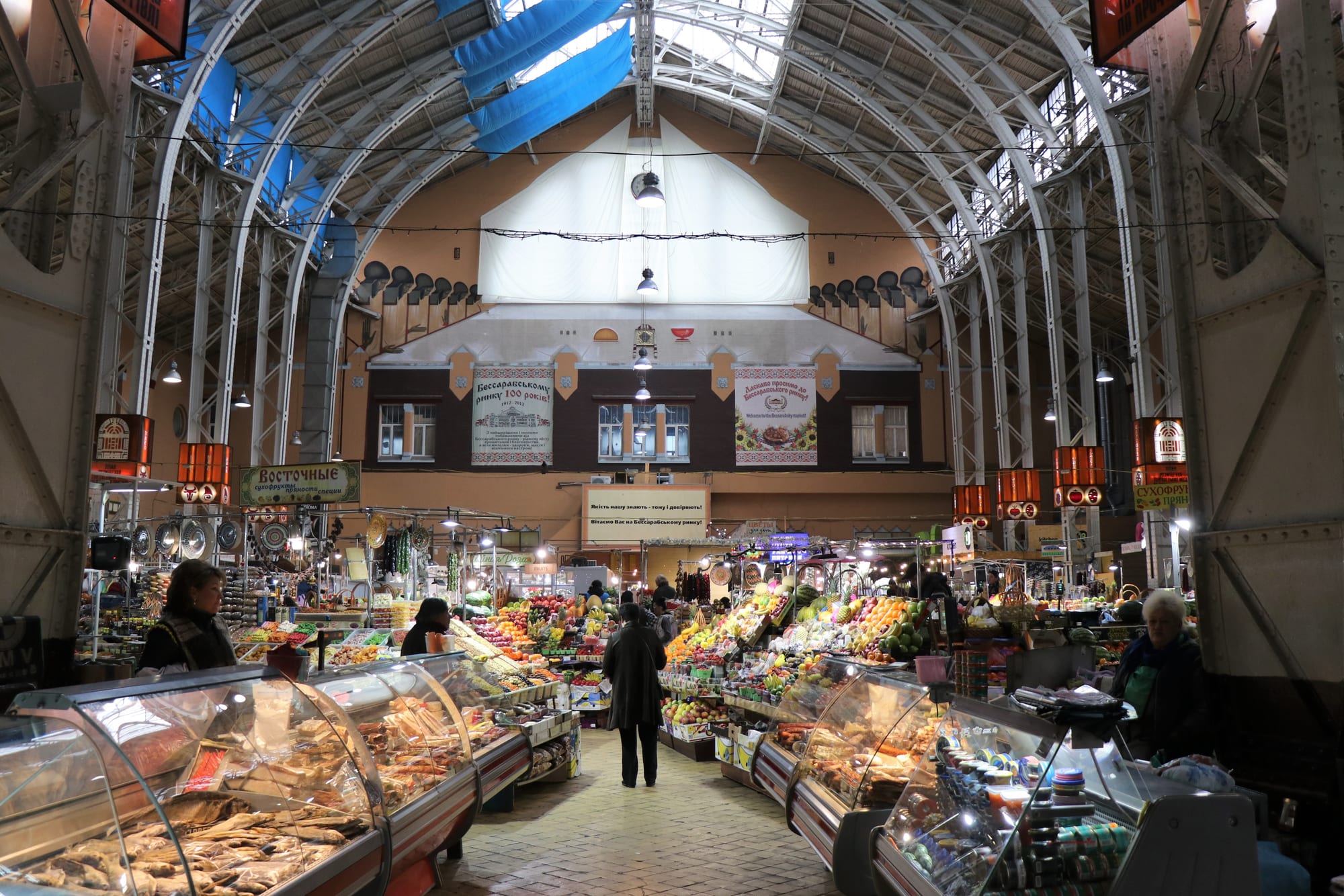 The colourful, well-stocked interior of Bessarabsky Market in Kyiv, with stallholders and shoppers among fruit, vegetables and deli counters under a high arched glass roof