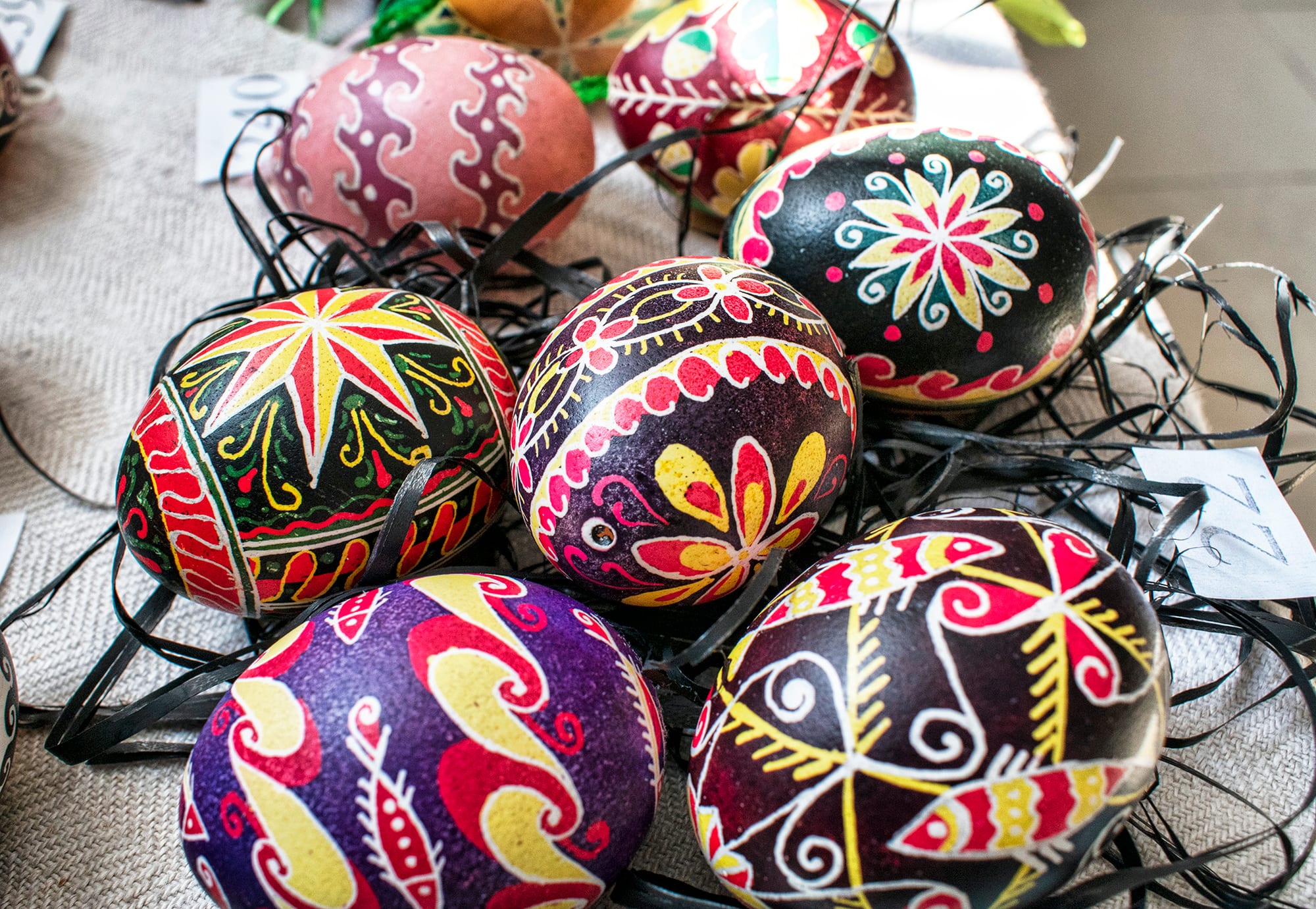 Seven eggs decorated with bold patterns of stars, flowers, spirals and waves in red, yellow, white, purple and black, resting on a bed of dark shredded paper