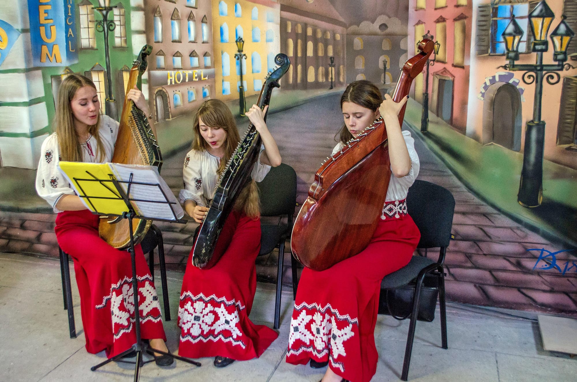 Three young women in white embroidered blouses and long red skirts with white patterns sit playing large many-stringed banduras, one singing from sheet music on a stand, in front of a painted street mural
