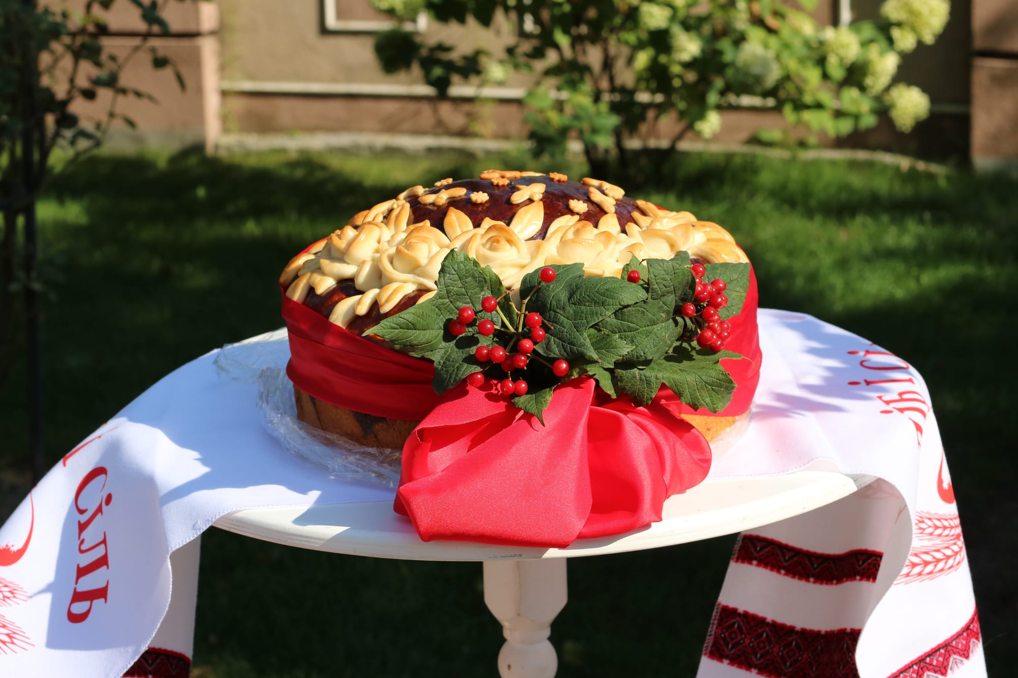 A large round loaf decorated with dough roses and wrapped in a red ribbon with green leaves and red berries, on a small white table draped with an embroidered cloth bearing red lettering