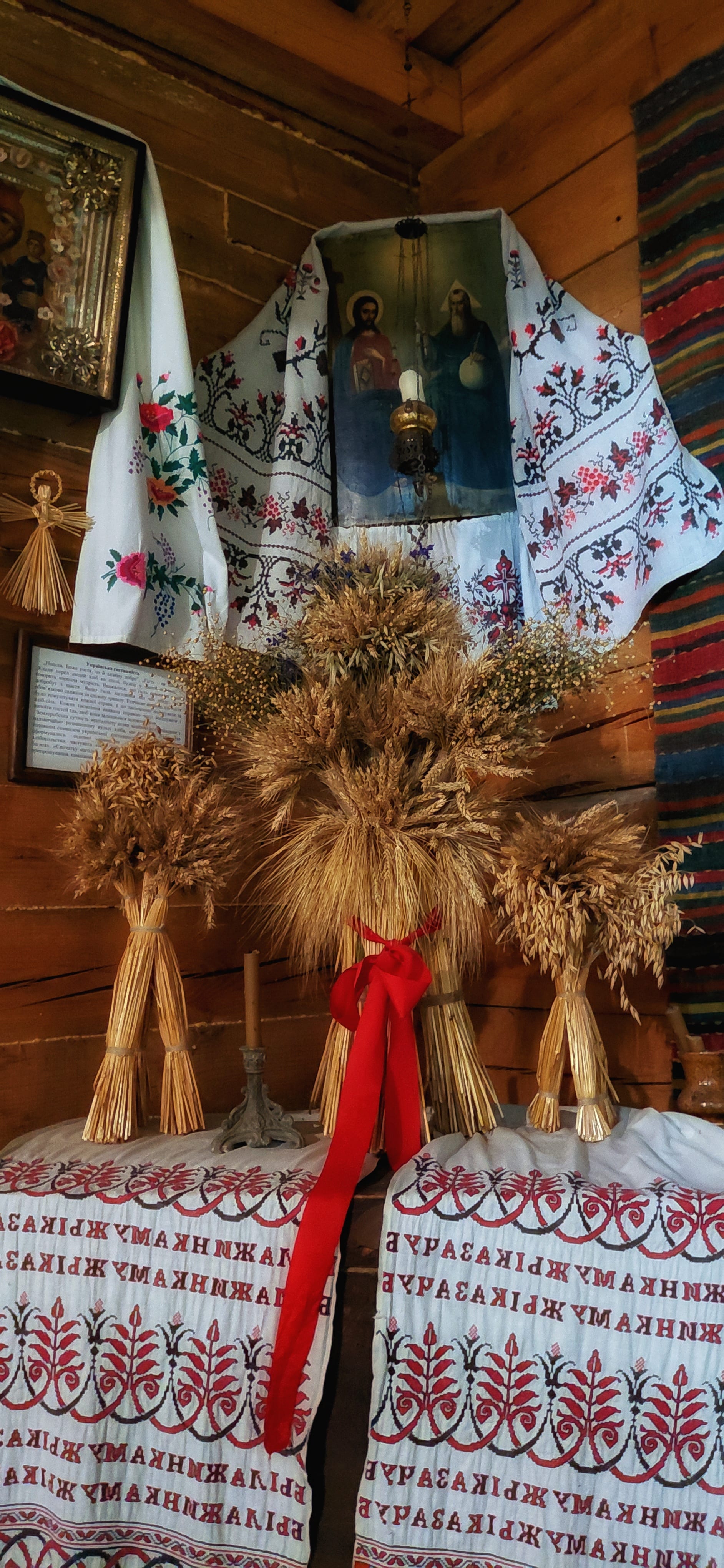 Sheaves of wheat and oats, the largest tied with a red ribbon, stand on embroidered cloths in the corner of a log-walled room beneath icons draped in embroidered towels
