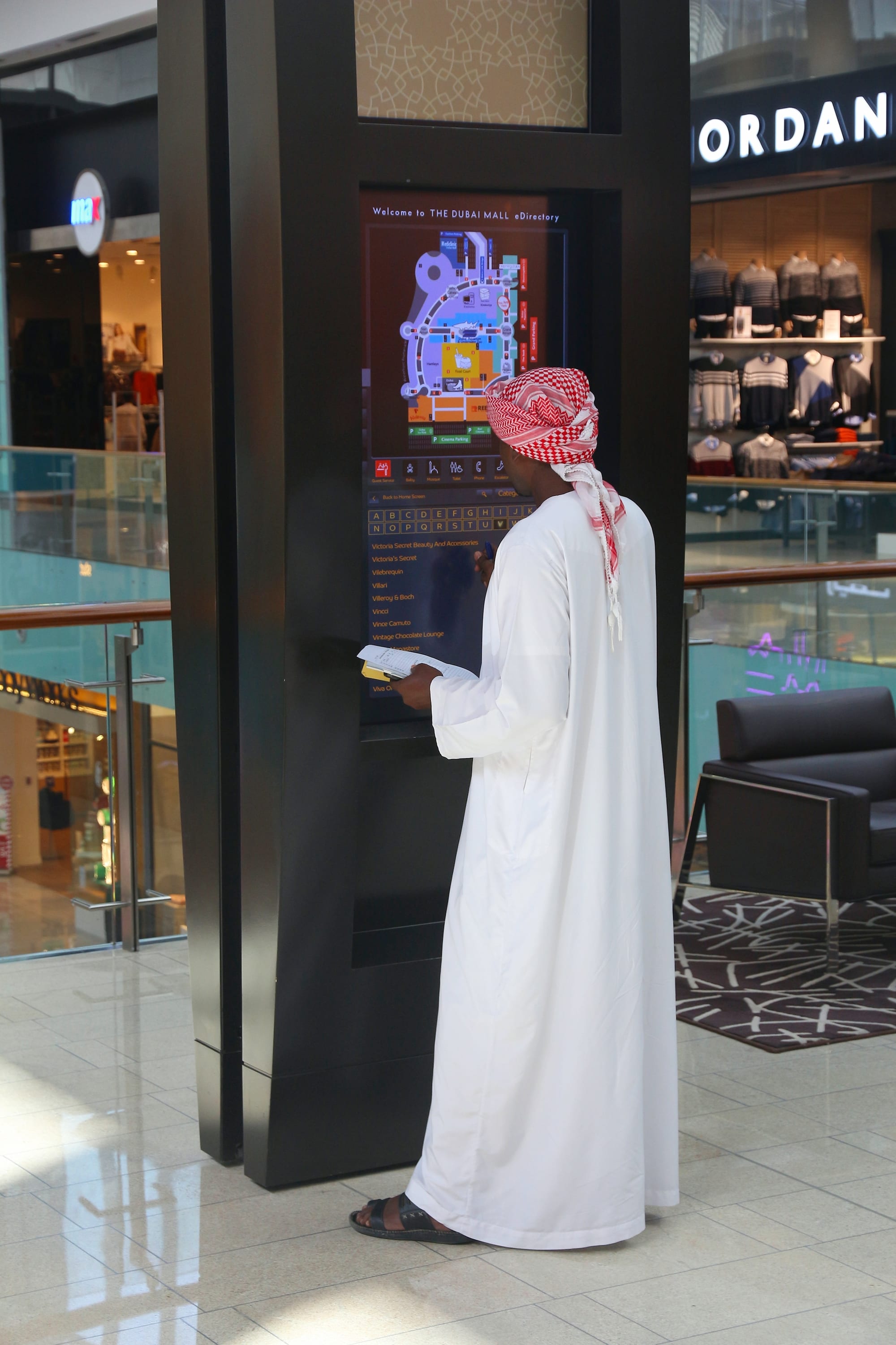 A man in a white kandura and red-and-white headscarf checking an electronic mall directory inside the Dubai Mall