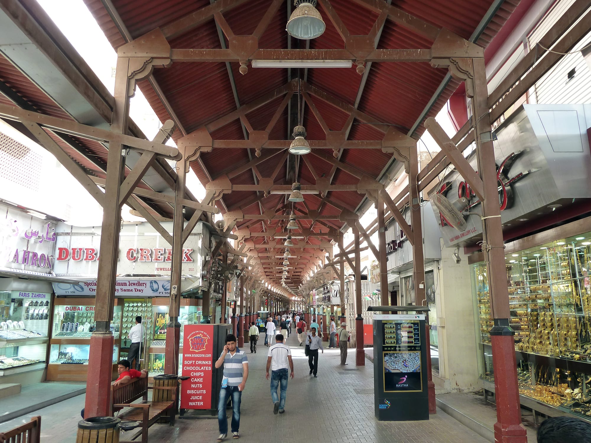 A covered market arcade in the Dubai Gold Souk, with jewellery shop windows full of gold on either side and shoppers walking through