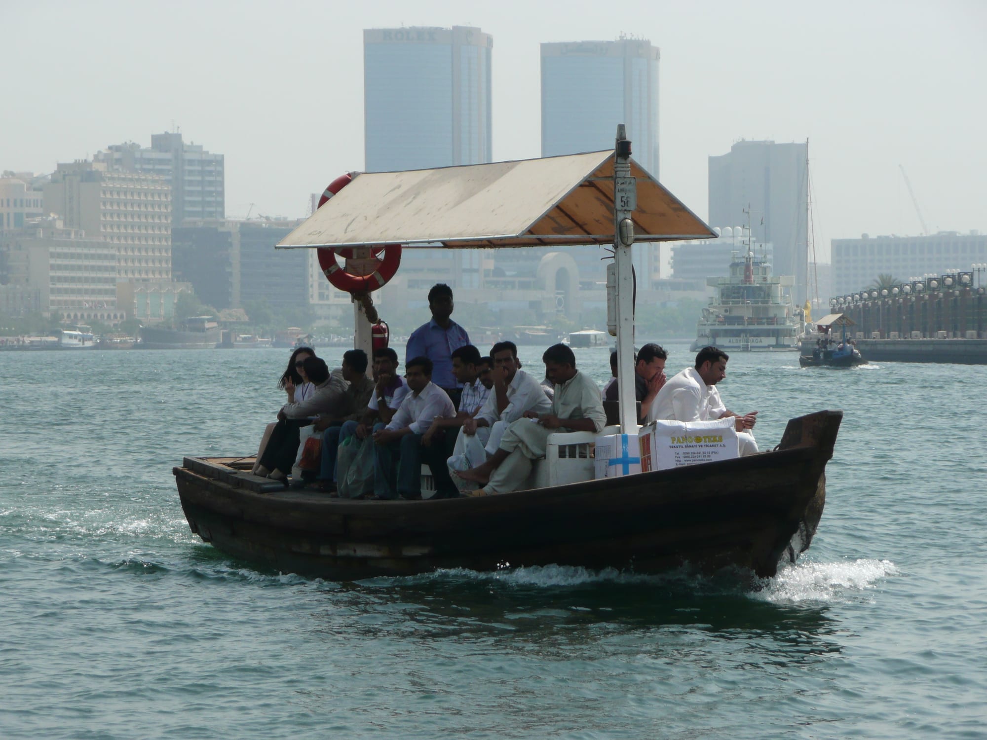 A wooden abra ferry crowded with passengers crossing Dubai Creek, with the city skyline behind