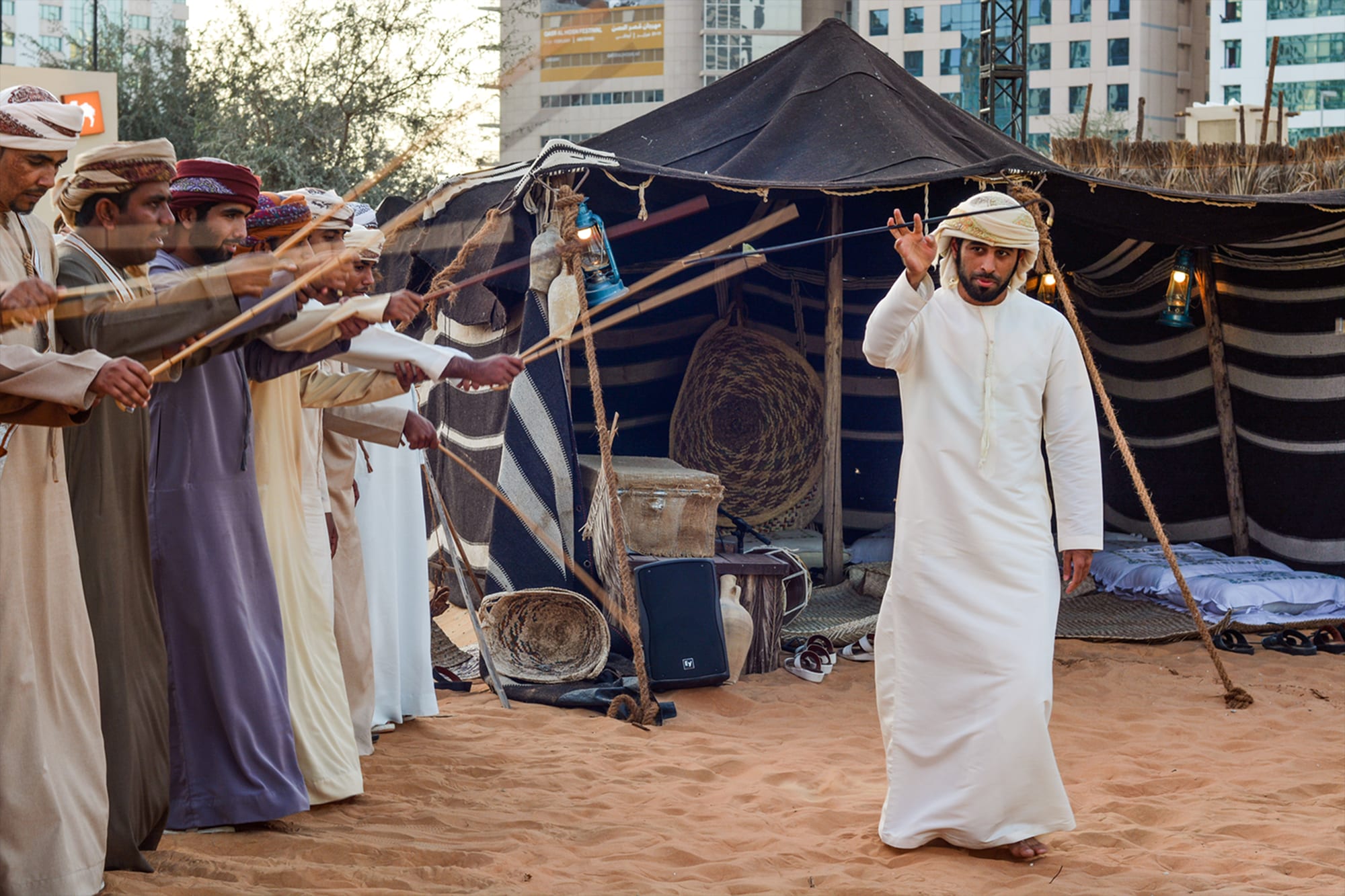 A row of men in long robes and head cloths hold thin canes aloft on the sand in front of a black tent, while a man in a white kandura stands before them with one hand raised