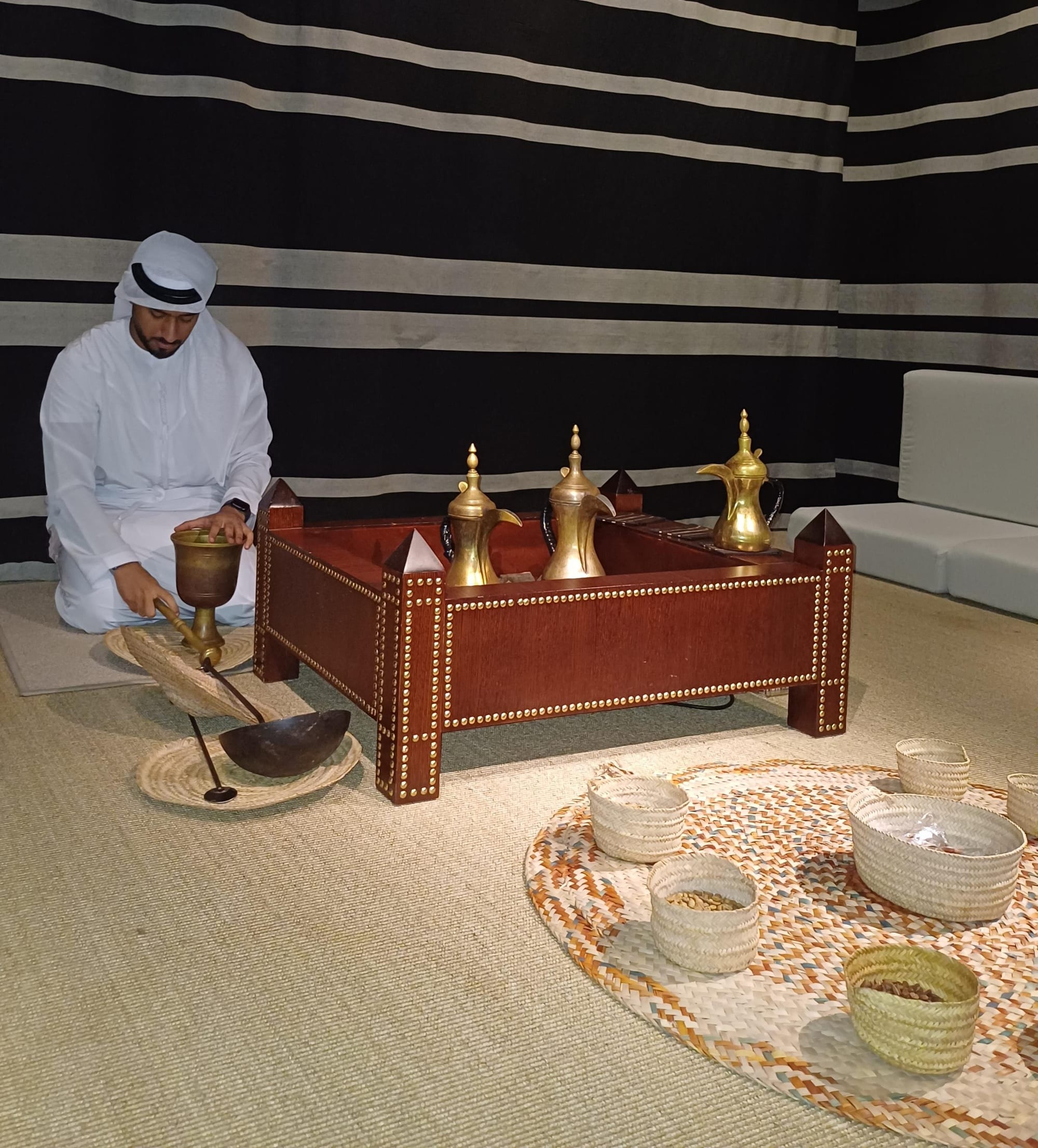 A man in a white kandura and head cloth kneels on a woven mat pounding coffee in a brass mortar beside a wooden hearth box holding three brass dallahs, with small woven baskets of coffee beans in the foreground