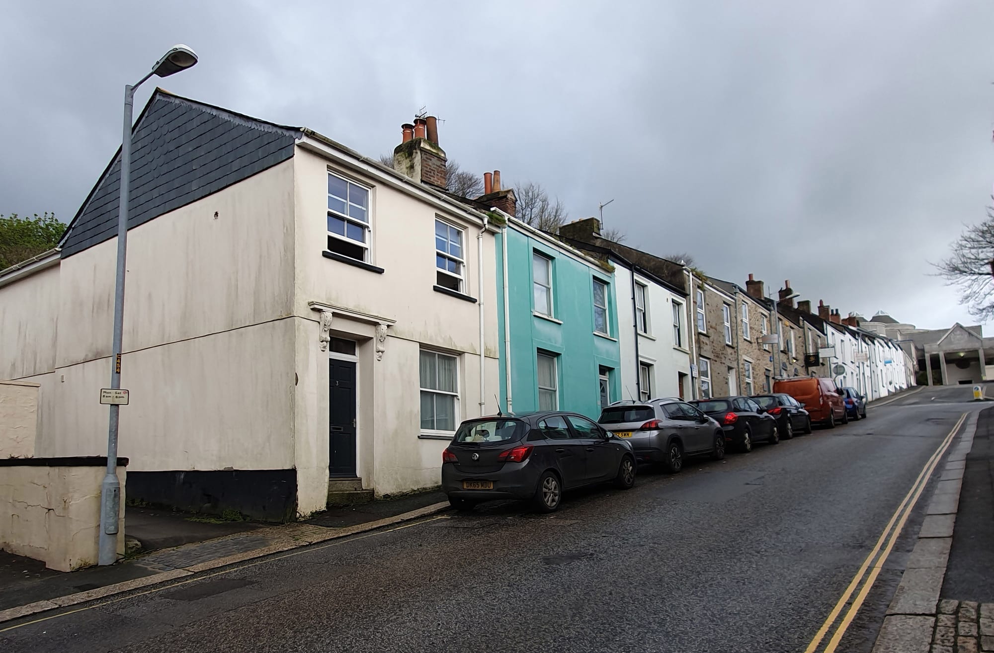 A row of terraced houses in different pastel colours on a sloping street in Truro, Cornwall, with cars parked along the kerb and a grey sky overhead