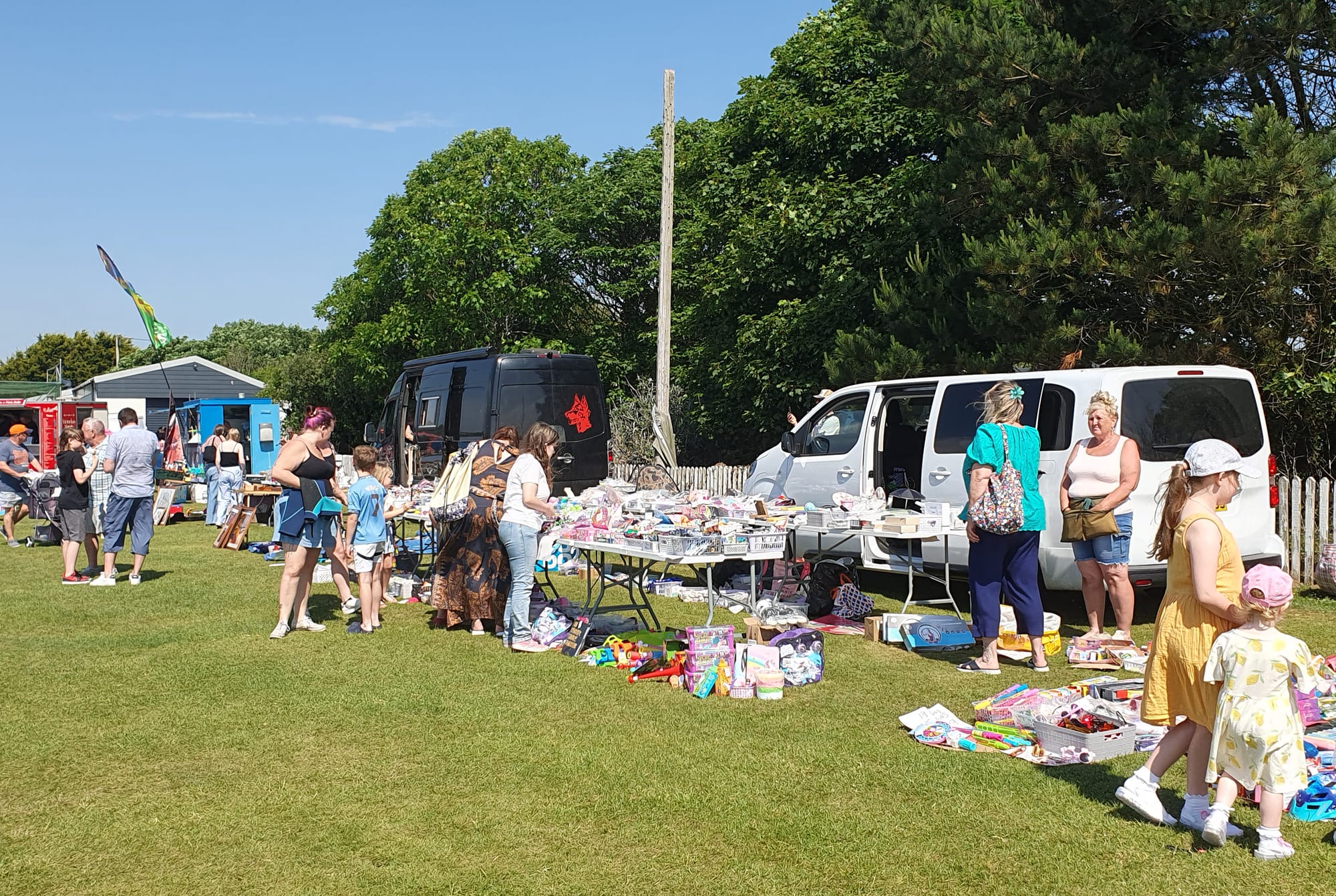 Two vans with their boots open and tables of secondhand goods laid out on a grass field at a car boot sale, with shoppers browsing under a clear blue sky