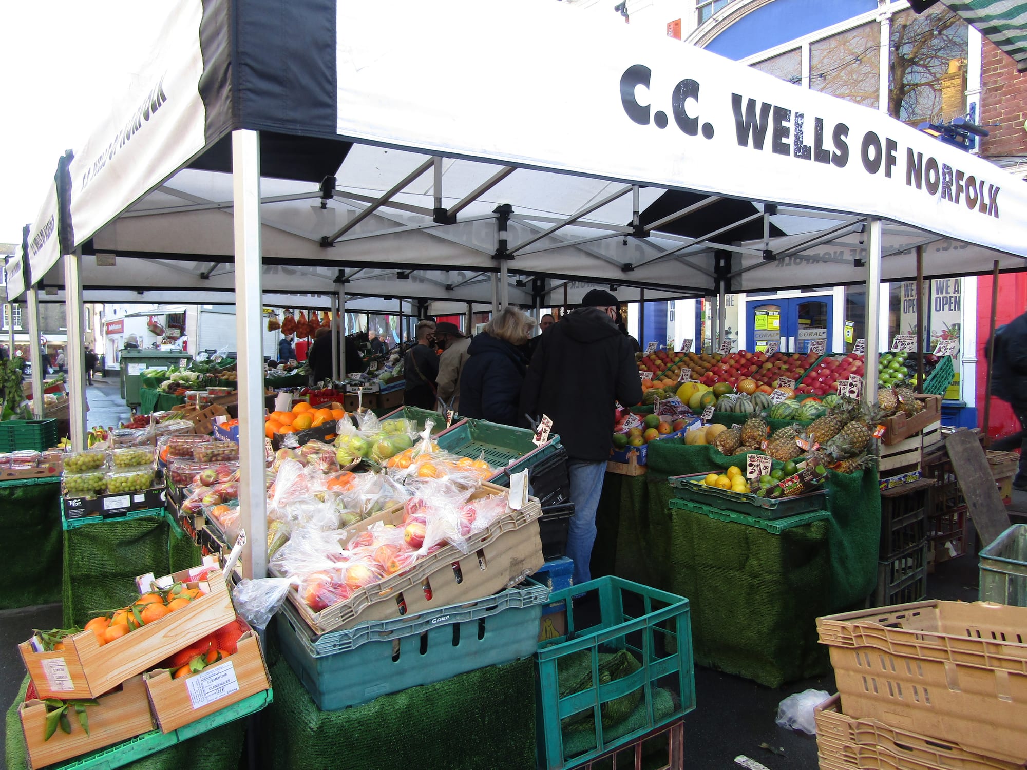 A crowded outdoor fruit and vegetable stall piled with apples, oranges, pineapples and melons under a market awning, with a shopper browsing