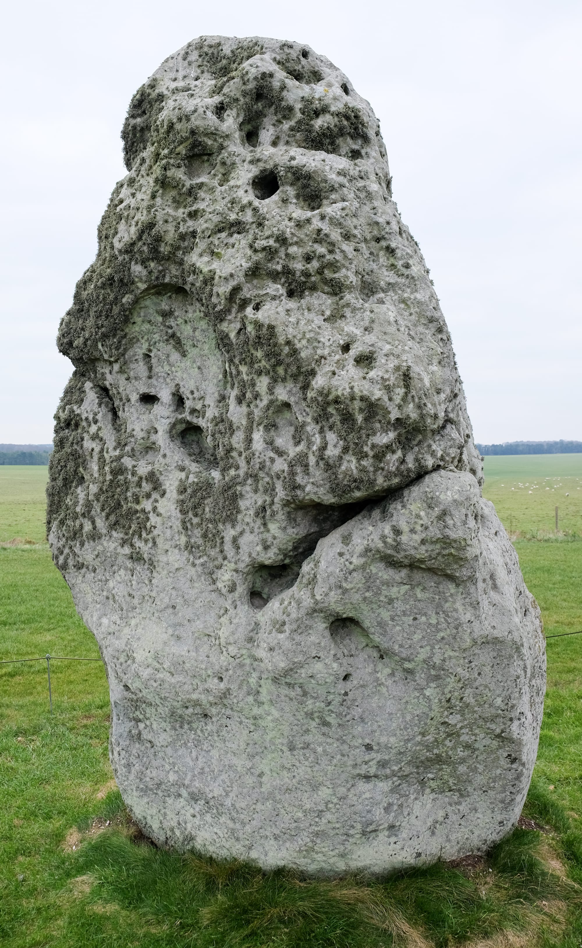 A single ancient standing stone in a field with the main stone circle visible behind it