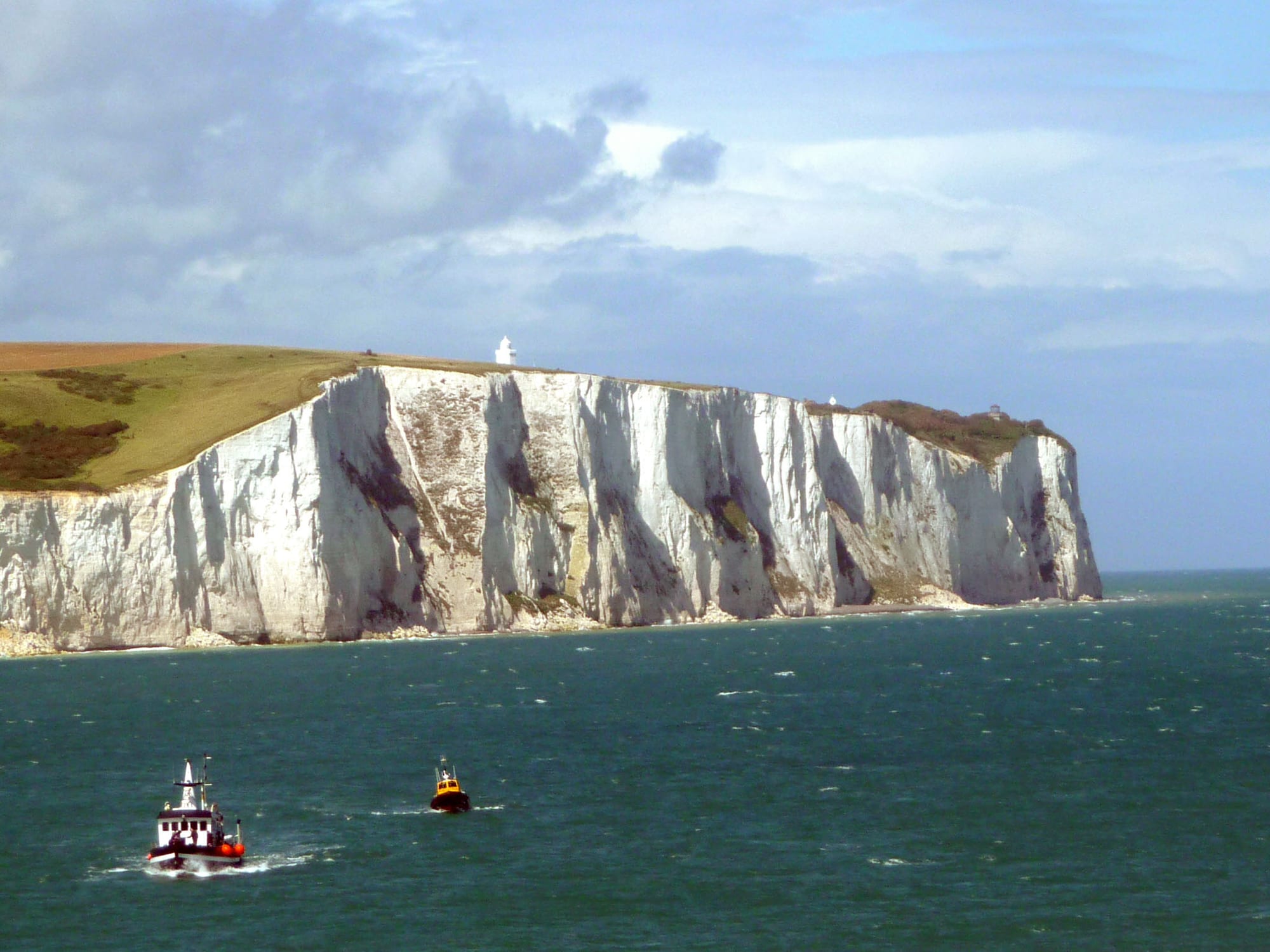 Sheer white chalk cliffs rising above the sea, with a lighthouse on top and small boats passing below