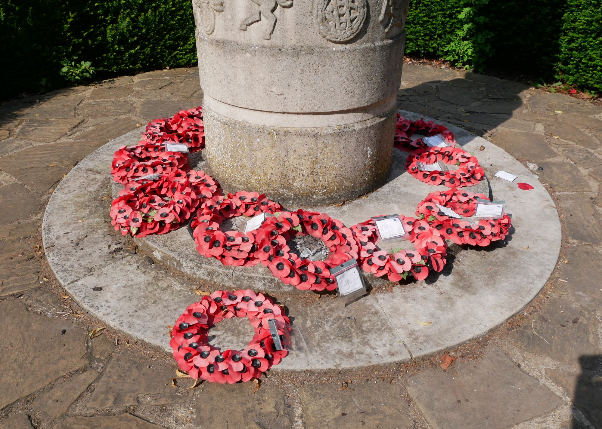 Wreaths of red paper poppies, each with a small card attached, laid in a ring around the stone base of a war memorial on a paved circle