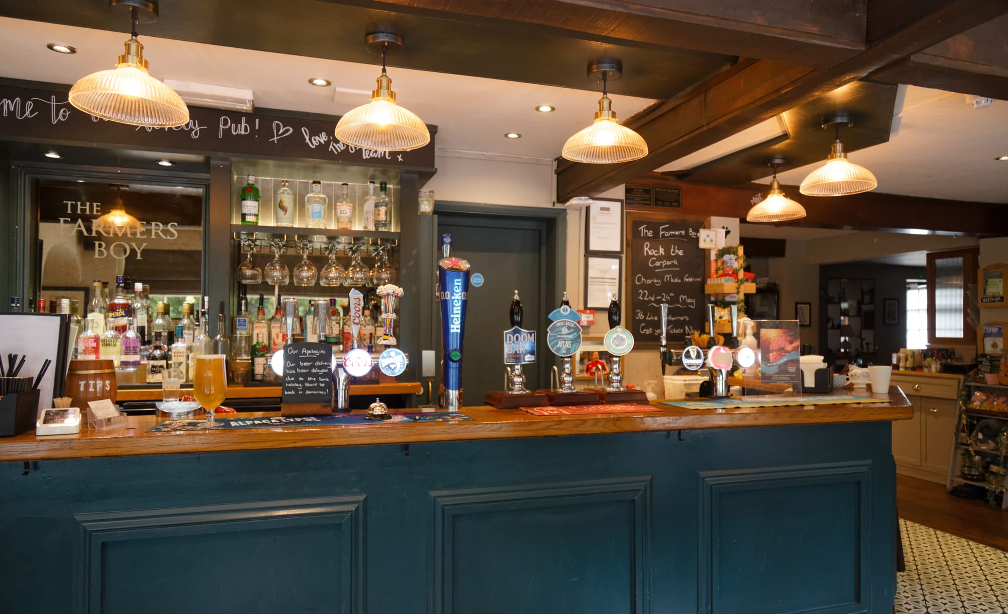 A wooden pub bar with a row of beer hand pumps and taps along the counter, a pint of lager resting on it, shelves of spirits behind and glass lamps hanging from a beamed ceiling