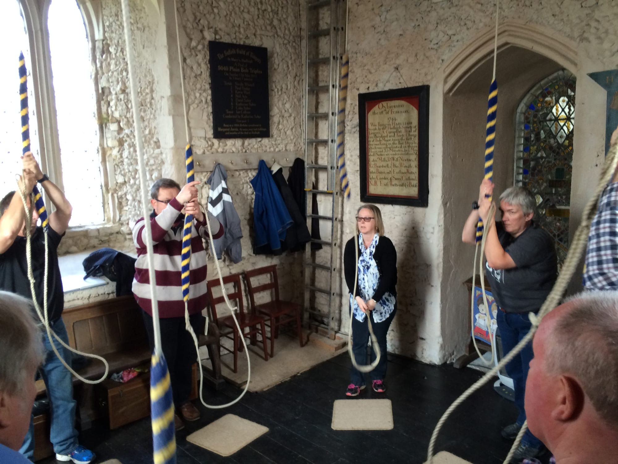 Several people stand in a circle in a stone-walled ringing chamber, each holding a long bell rope with a striped blue and yellow woolly grip, beneath framed boards recording past peals