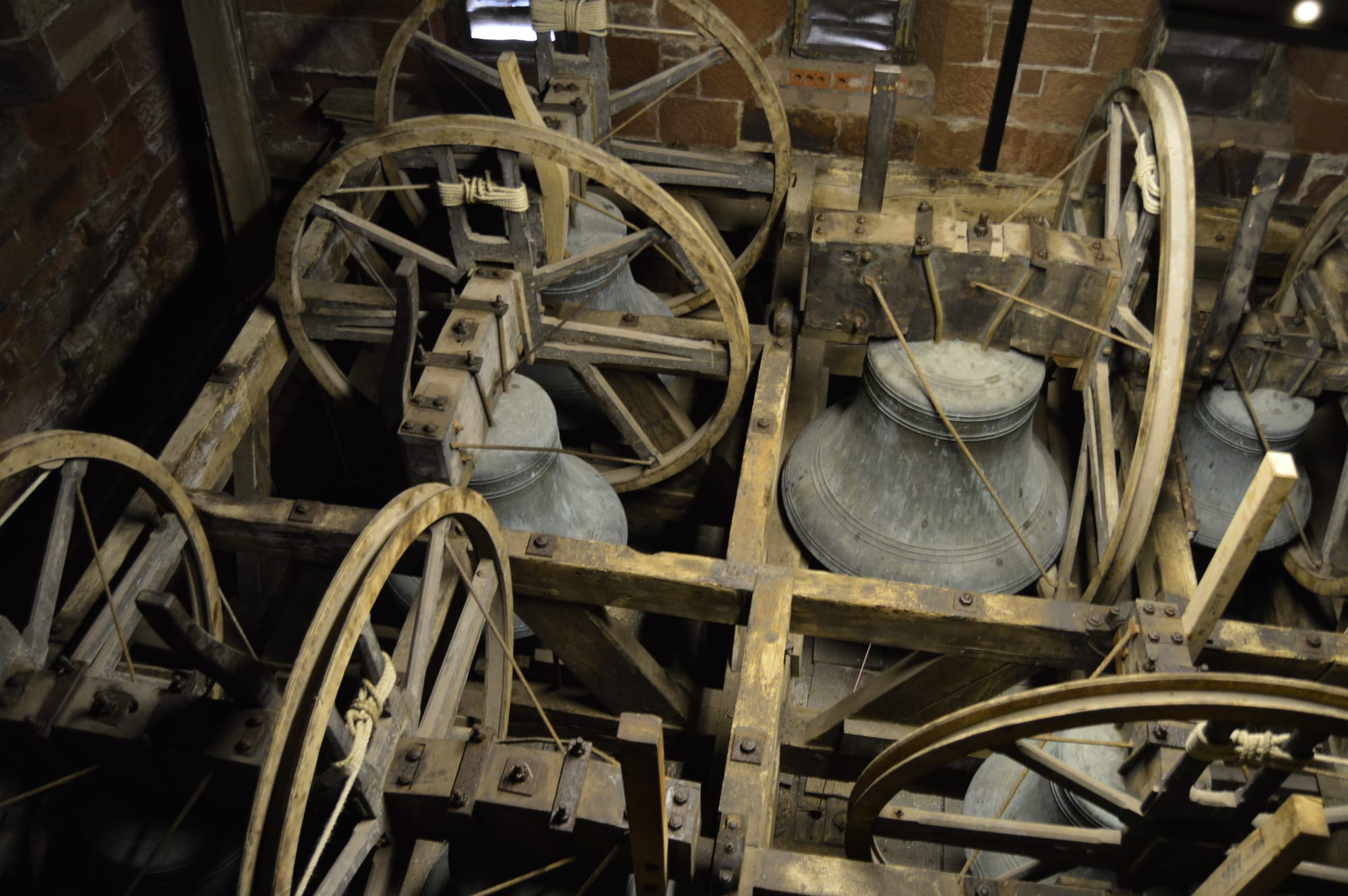 Large bronze church bells hanging mouth-down in a heavy wooden frame inside a brick tower, each mounted beside a big wooden wheel with a rope running around its rim