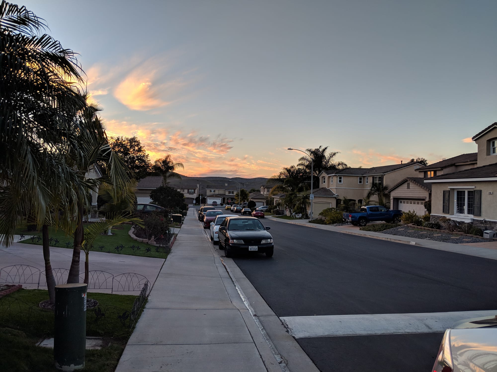 A wide suburban street at sunset lined with parked cars, palm trees and single-family houses with garages