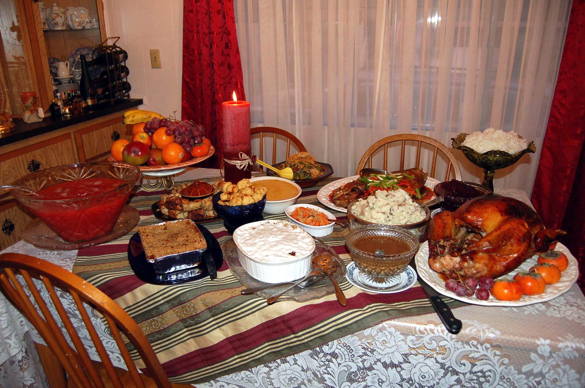 A dining table with a striped runner laid with a roast turkey, bowls of mashed potato, gravy and rice, a fruit bowl and a lit red candle