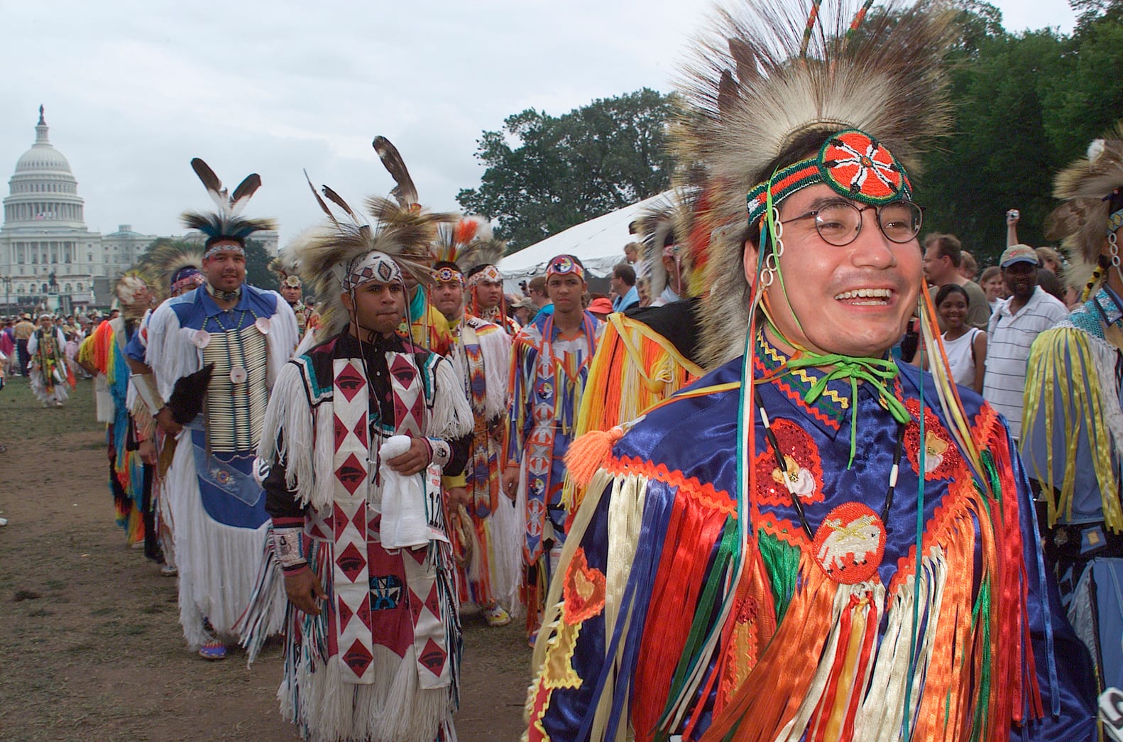 Native American dancers in feathered headdresses and brightly coloured fringed regalia walk in procession across a grassy field, with the white dome of the US Capitol in the background and spectators to the side