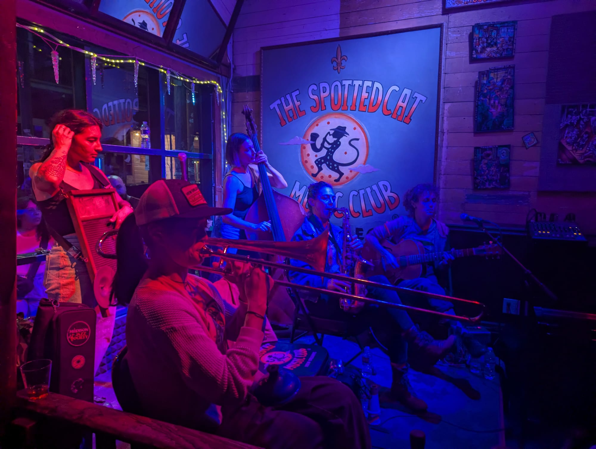 A jazz band with a trombone, washboard, double bass, clarinet and guitar plays on a small stage under a sign reading The Spotted Cat Music Club