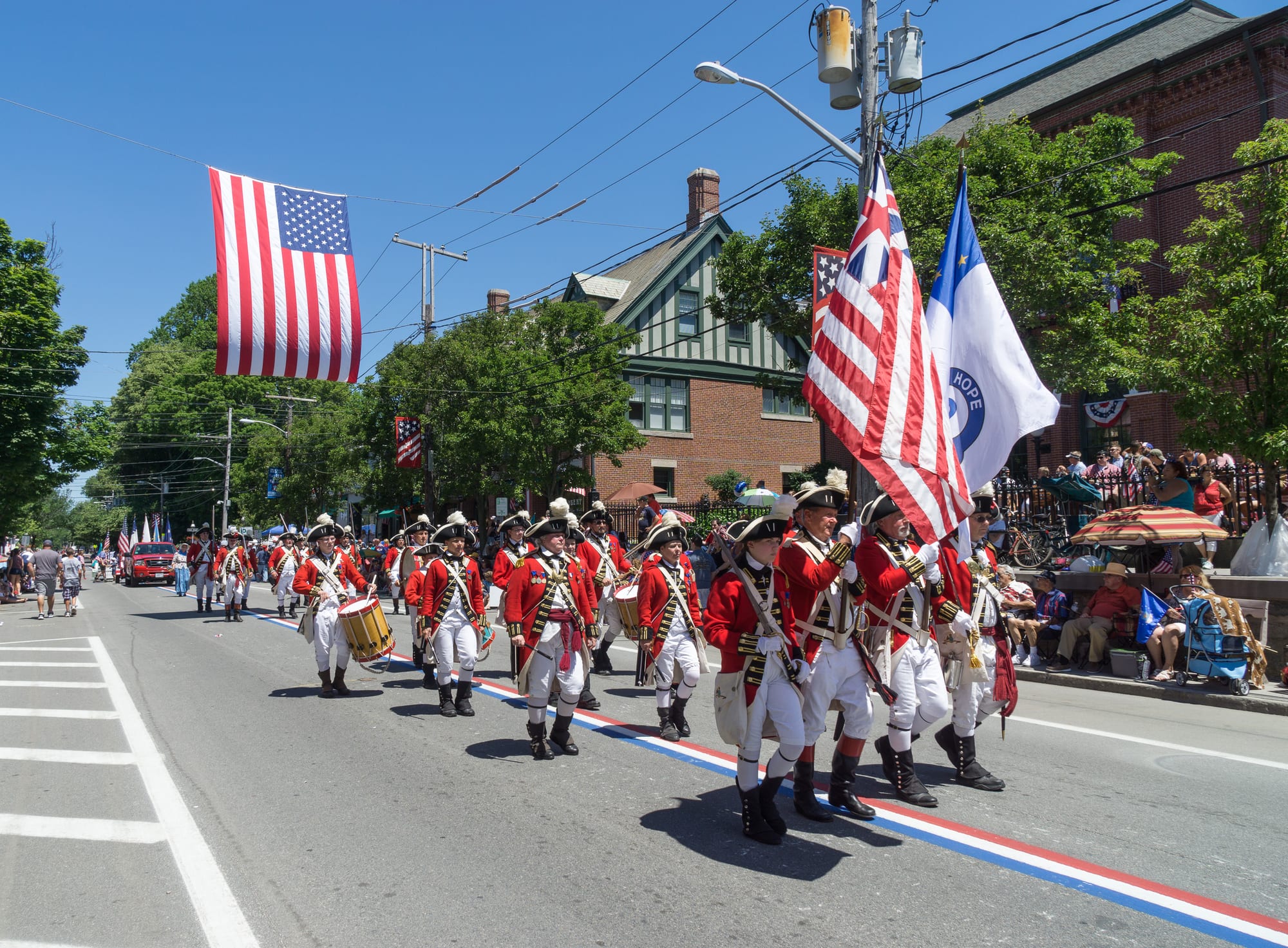 Marchers in red coats, white breeches and tricorn hats carry drums and flags down a street lined with spectators, under a large US flag strung across the road