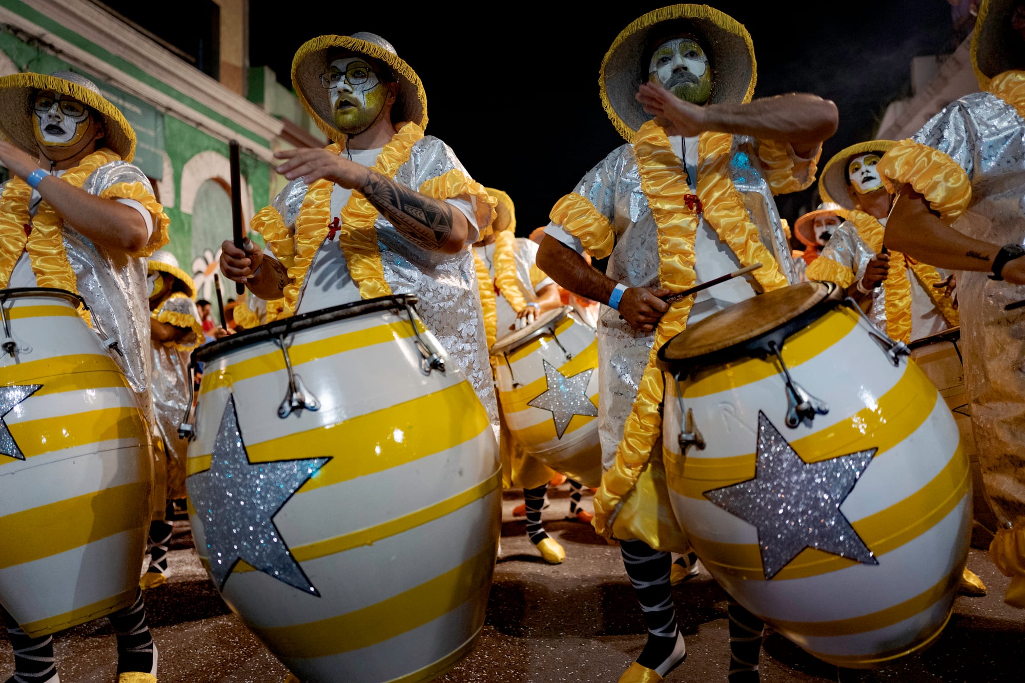 Drummers with white-painted faces, straw hats and shiny silver and yellow costumes play large barrel-shaped drums painted with yellow stripes and silver stars at night