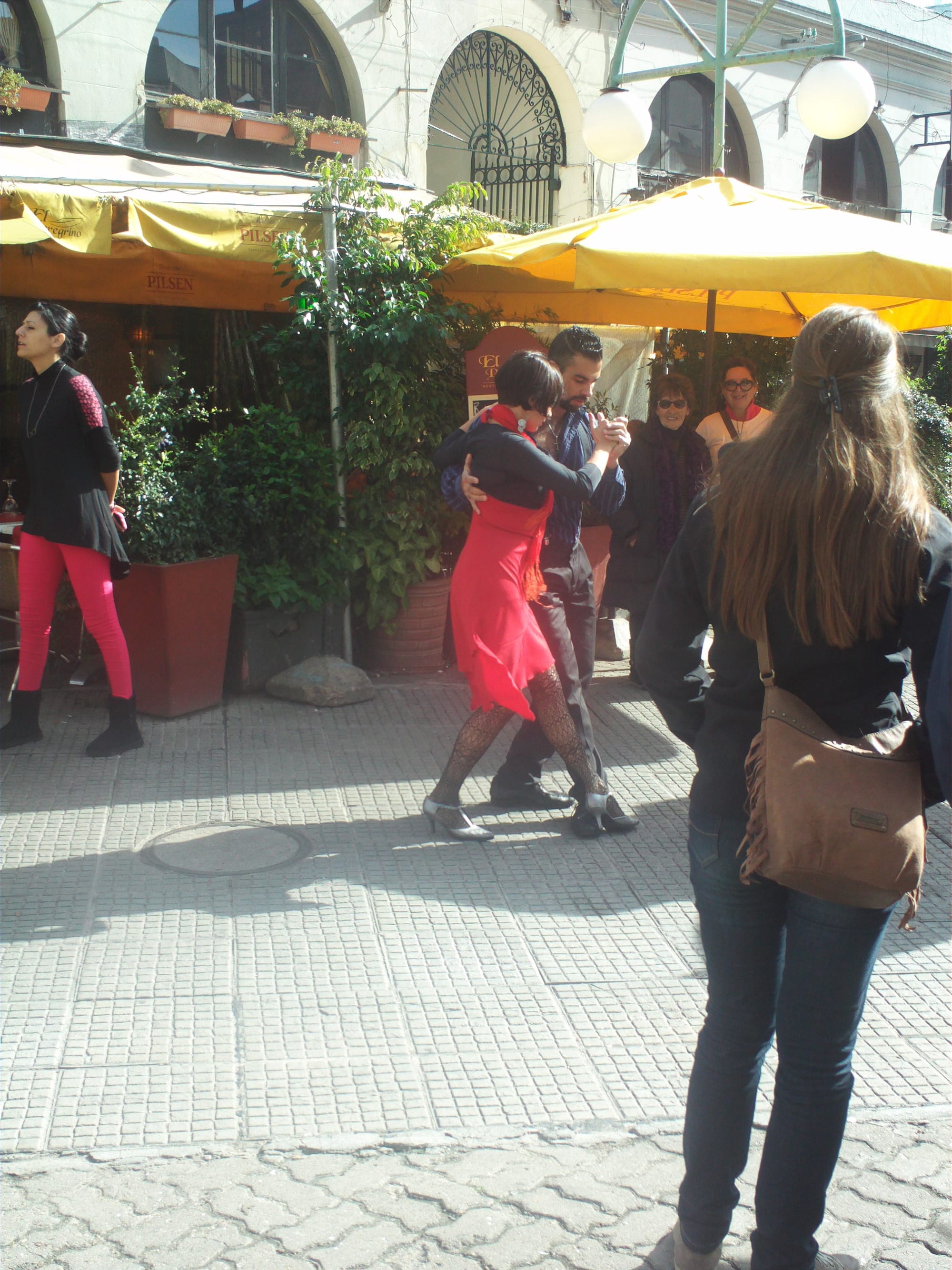 A couple in a close embrace dance tango on a paved pedestrian street outside a café with yellow umbrellas, watched by passers-by