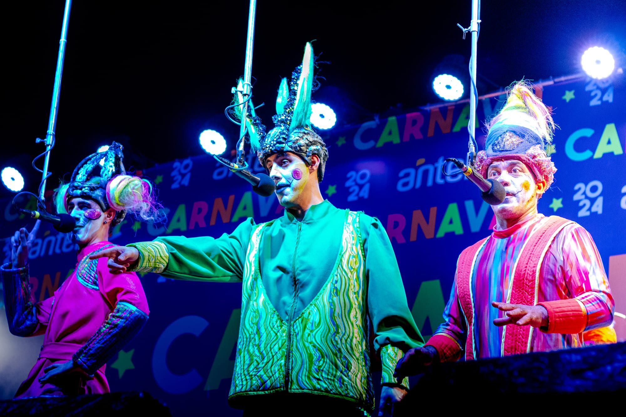 Three murga singers in brightly coloured costumes, tall decorated headdresses and painted faces sing into overhead microphones on a stage lit with blue lights and a Carnaval 2024 backdrop